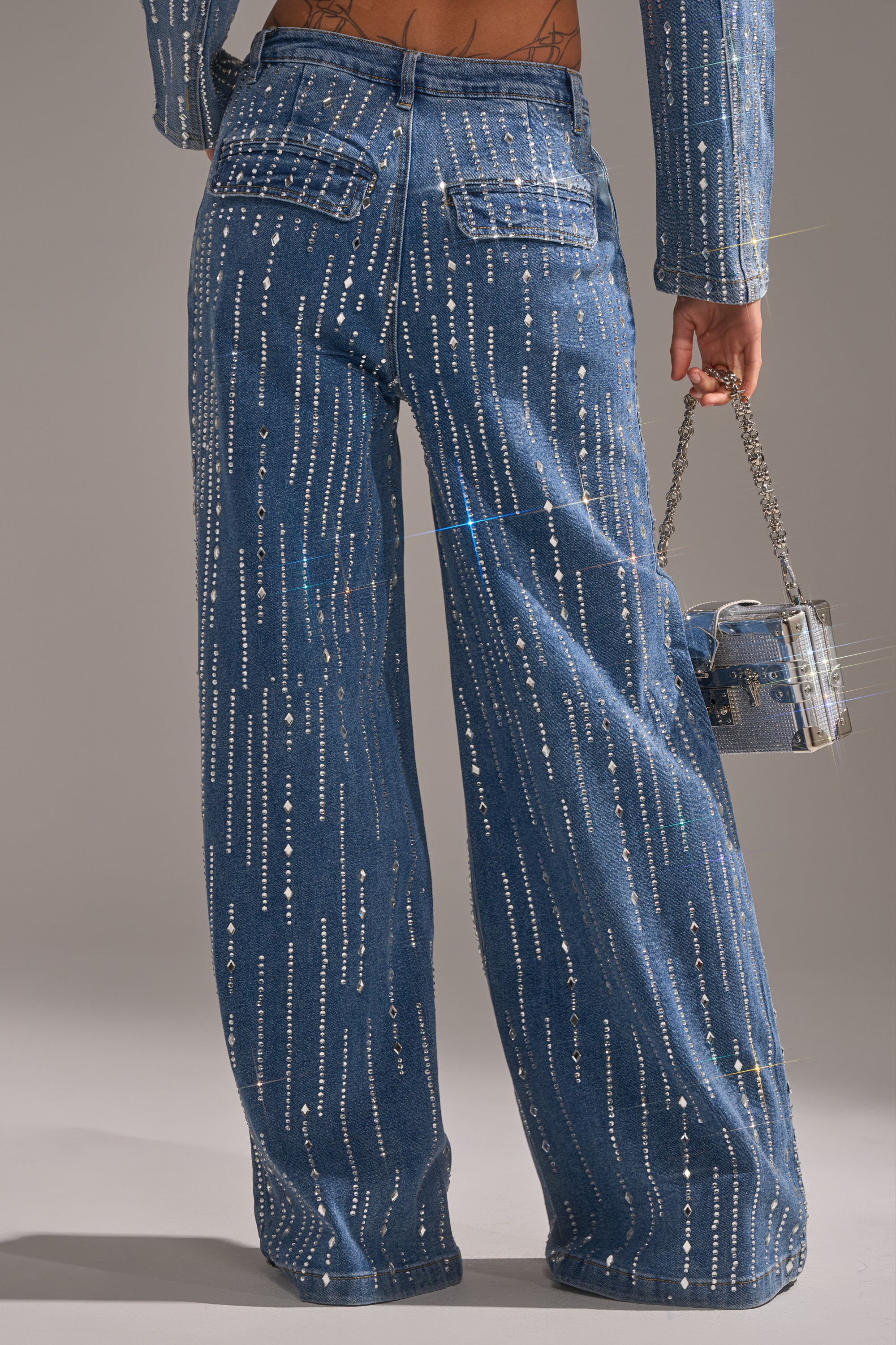 A person wears the NEVER COME DOWN RHINESTONE DENIM—blue high-rise, wide-leg jeans with silver sequin stripes—paired with a small chain-strap matching bag. The image is taken from behind, highlighting the jean silhouette and bag.