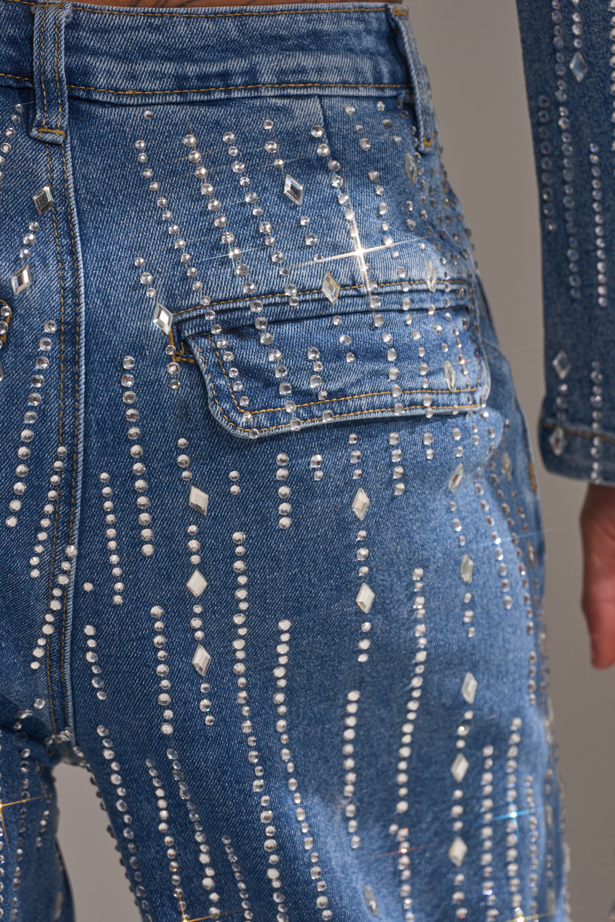 Close-up of the NEVER COME DOWN RHINESTONE DENIM’s back pocket, showcasing high-rise wide-leg jeans adorned with rows of sparkling rhinestones and metallic studs in assorted shapes for a dazzling, patterned effect.