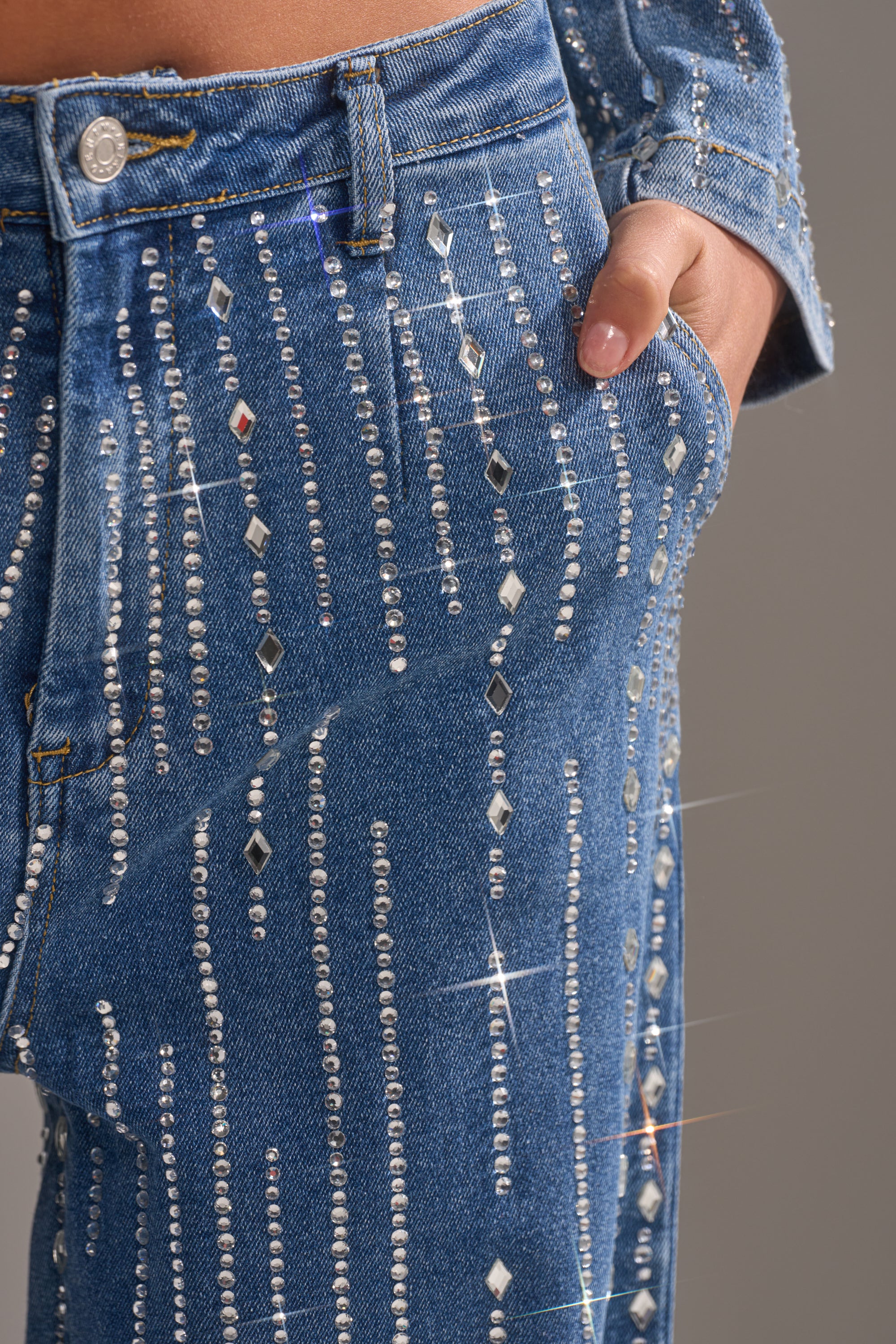 Close-up of a person wearing NEVER COME DOWN RHINESTONE DENIM: blue high-rise jeans and matching jacket, both featuring vertical rows of sparkling rhinestones. A hand in the pocket highlights the eye-catching design.