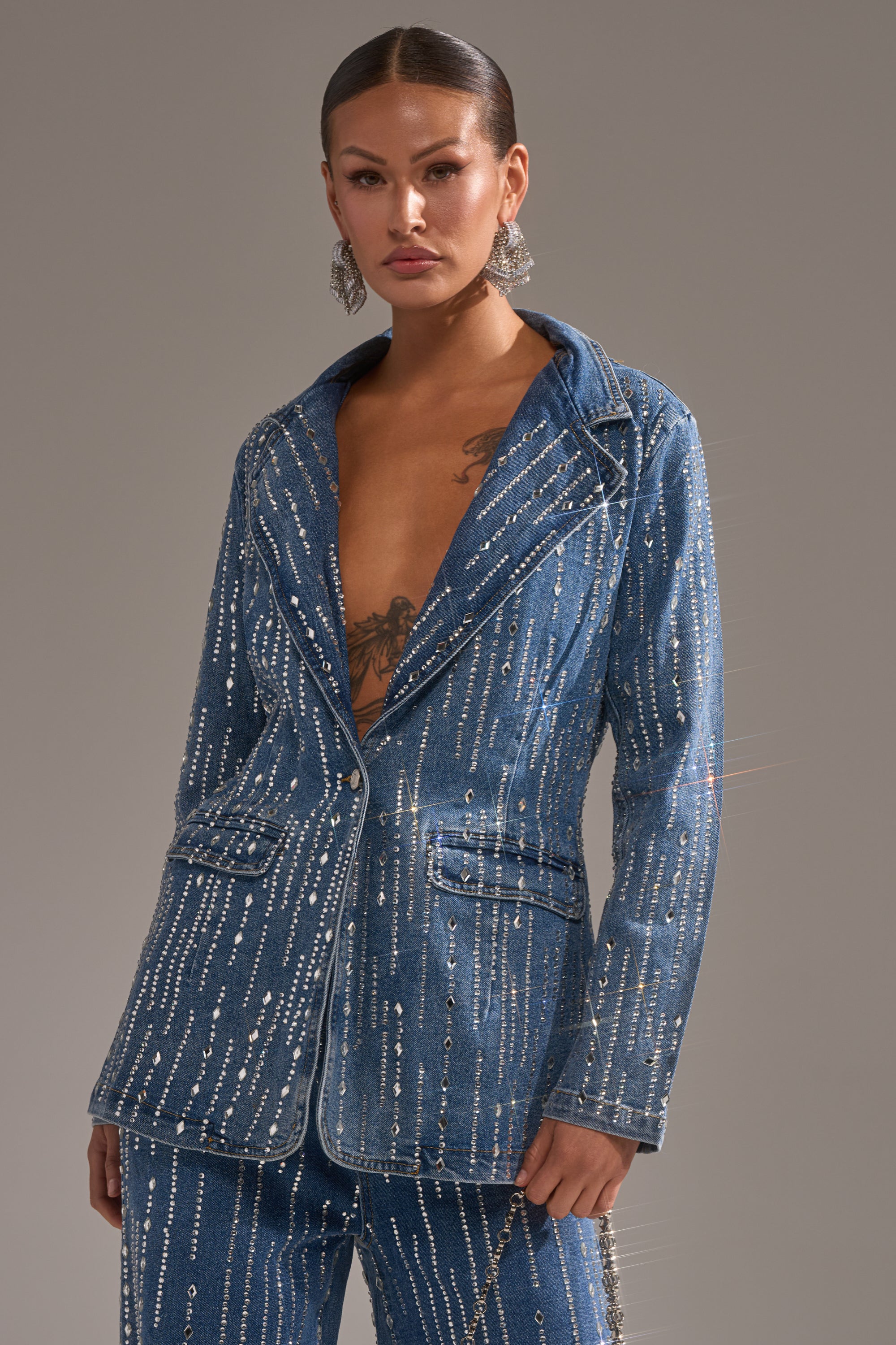 Wearing the NEVER COME DOWN RHINESTONE DENIM BLAZER, a person with slicked-back hair and visible chest tattoos poses confidently against a neutral background in fitted, blue denim adorned with sparkling rhinestone accents.