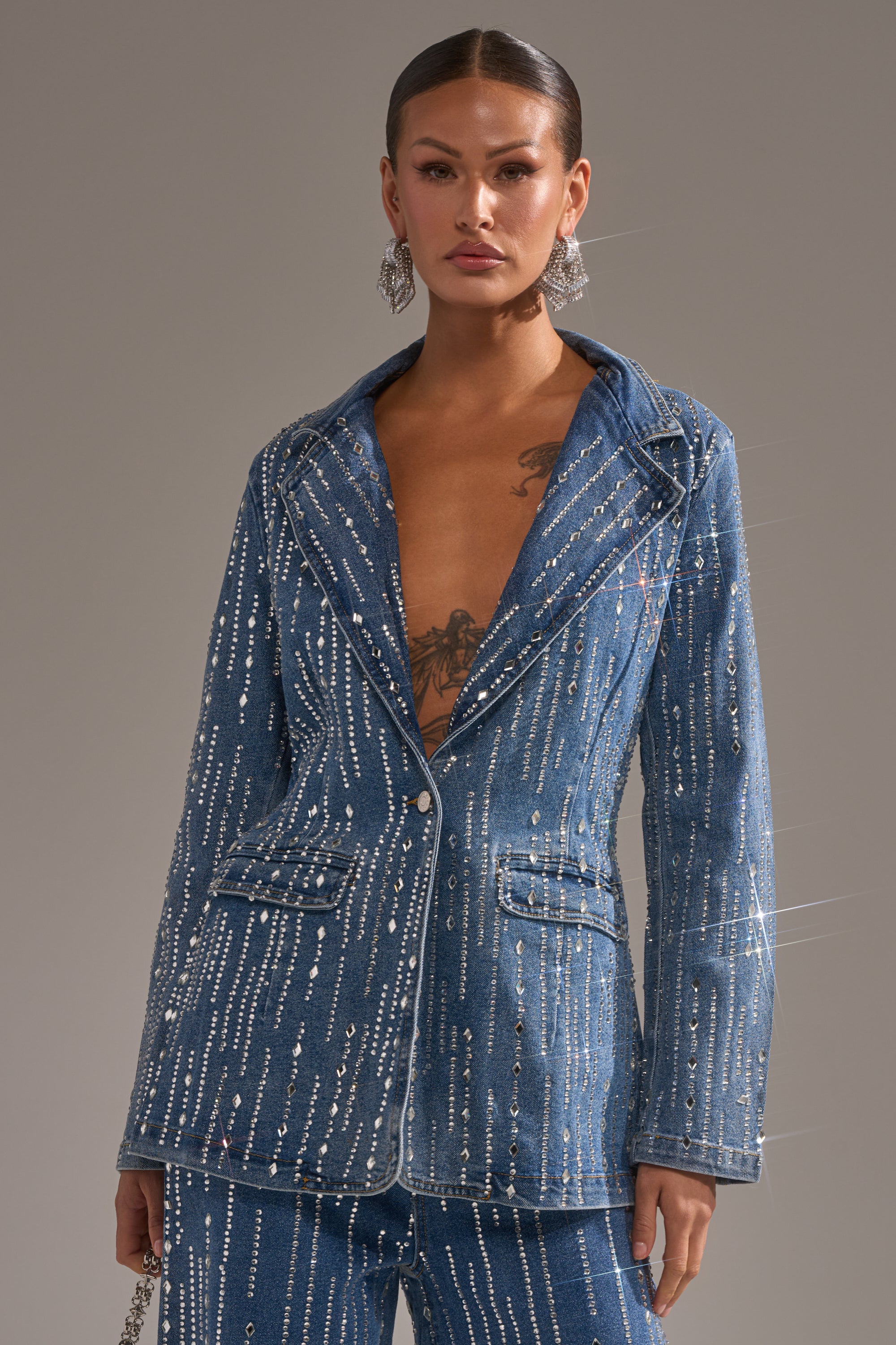 A person models the NEVER COME DOWN RHINESTONE DENIM BLAZER with sparkling rhinestones and matching pants, accessorized with bold dangling earrings, standing confidently against a plain background.