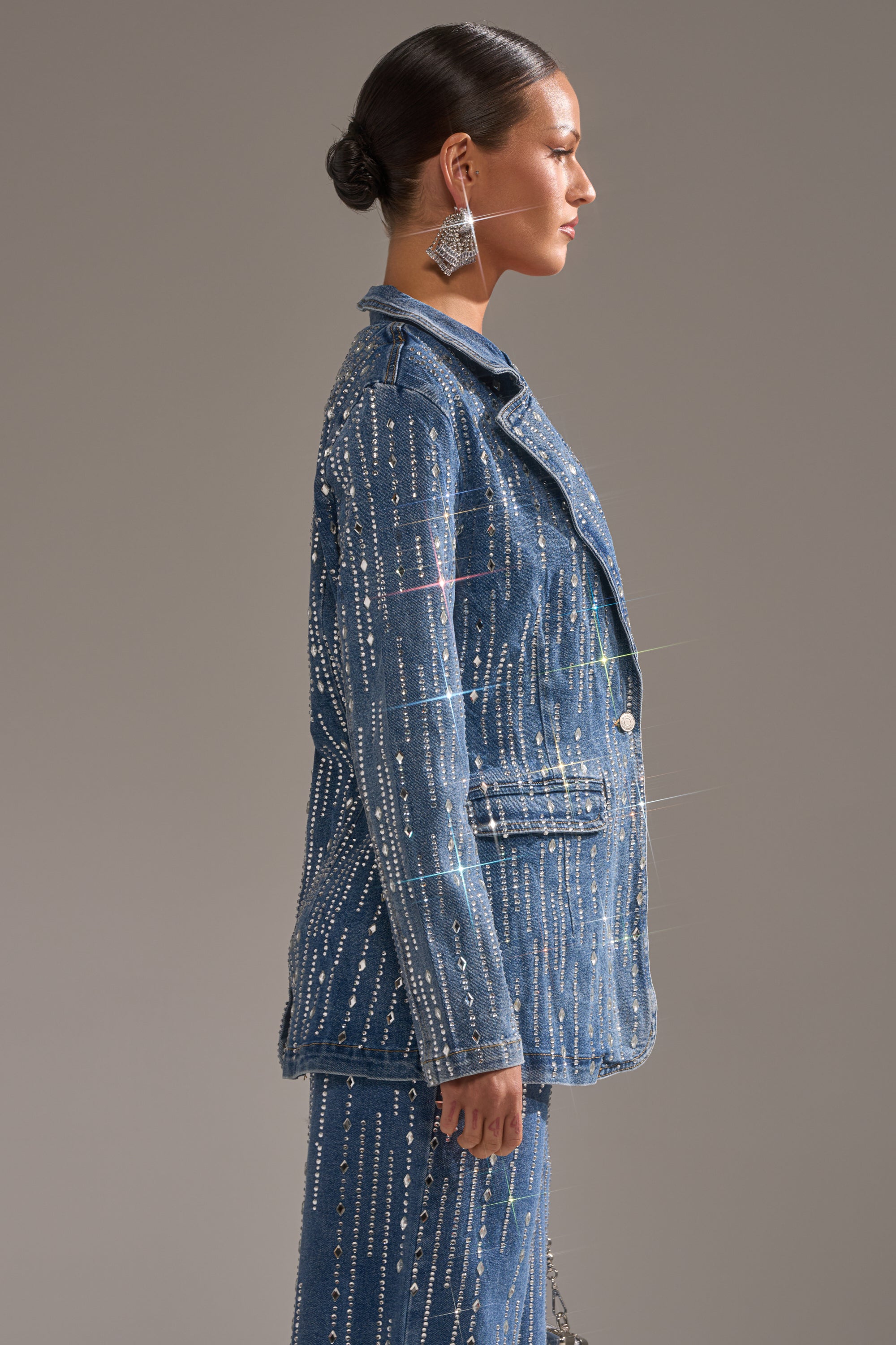 A woman stands in profile wearing the NEVER COME DOWN RHINESTONE DENIM BLAZER and matching pants with vertical rhinestone embellishments, large geometric earrings, and her dark hair styled in a sleek bun.