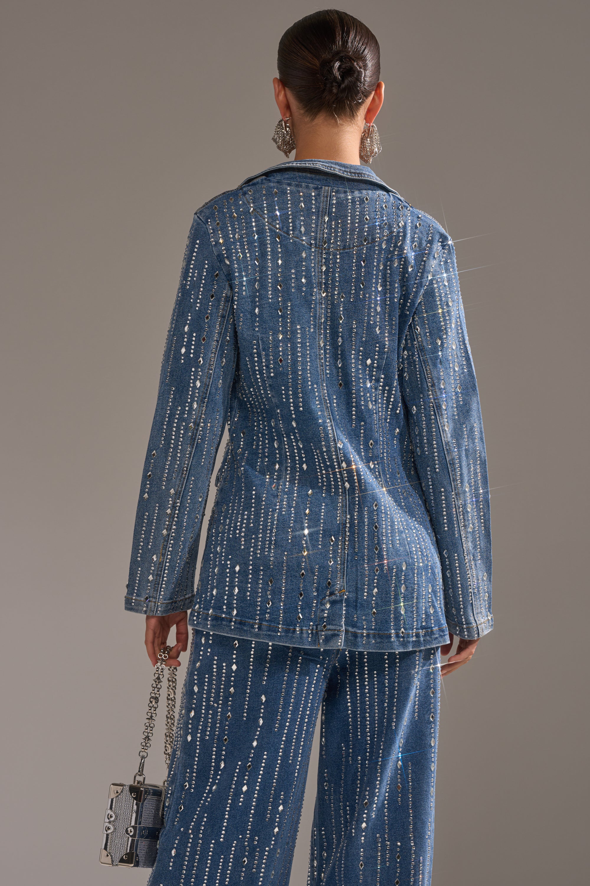 A woman with her hair in a sleek bun is seen from behind, wearing the NEVER COME DOWN RHINESTONE DENIM BLAZER with sparkling vertical embellishments. She carries a chain-strap handbag and wears large statement earrings.