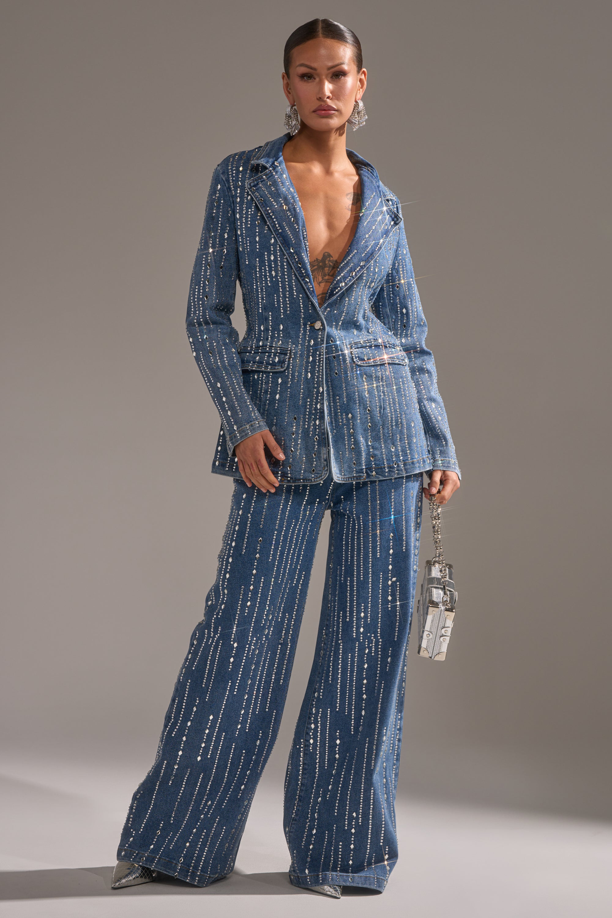 A woman models the NEVER COME DOWN RHINESTONE DENIM BLAZER with sequin details and a plunging neckline, pairing it with wide-leg pants, statement earrings, and a clear handbag against a plain gray background.