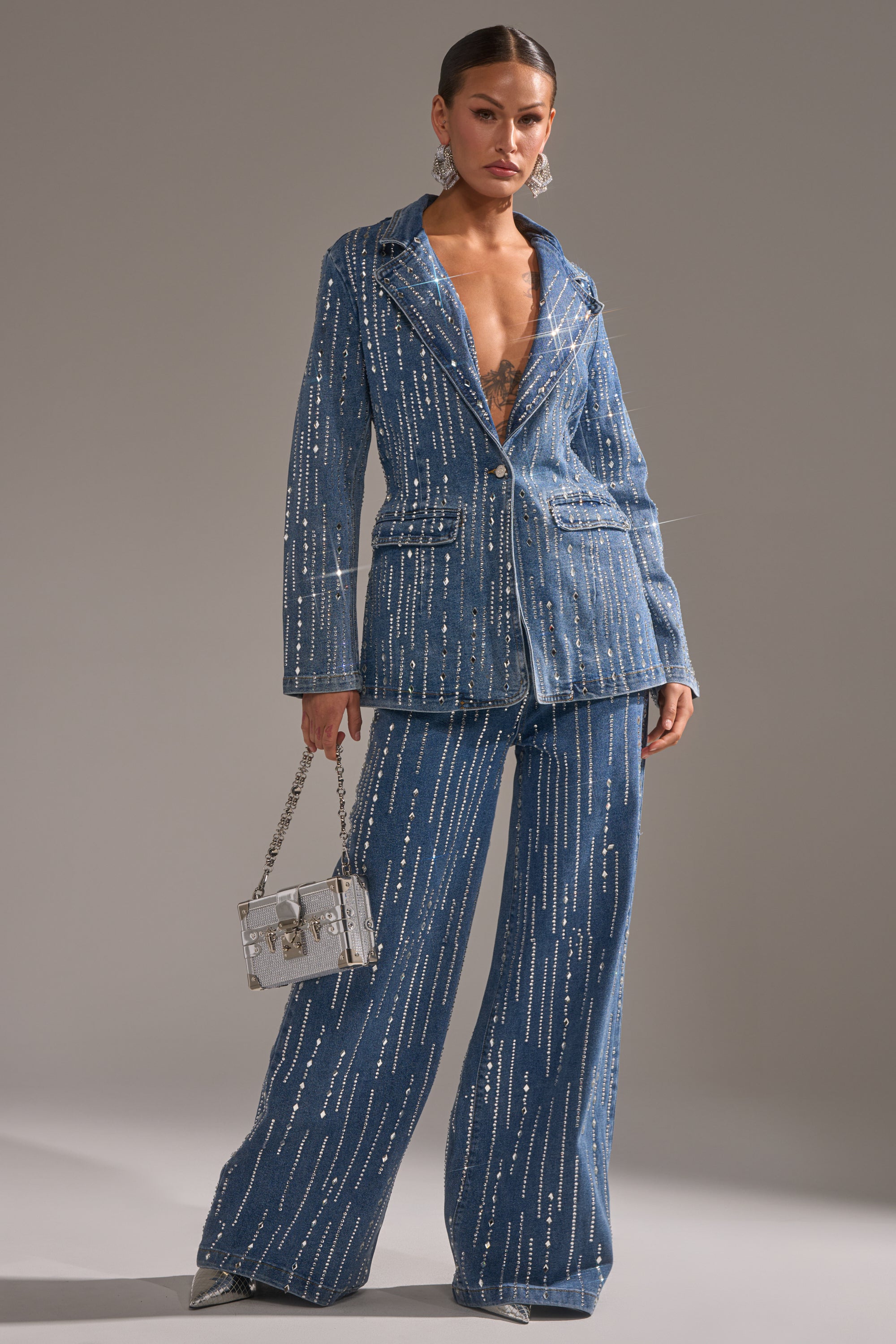 A woman stands confidently in the NEVER COME DOWN RHINESTONE DENIM BLAZER, featuring sparkly blue denim with silver vertical rhinestone embellishments. She holds a clear rectangular handbag and wears pointed shoes against a plain gray background.