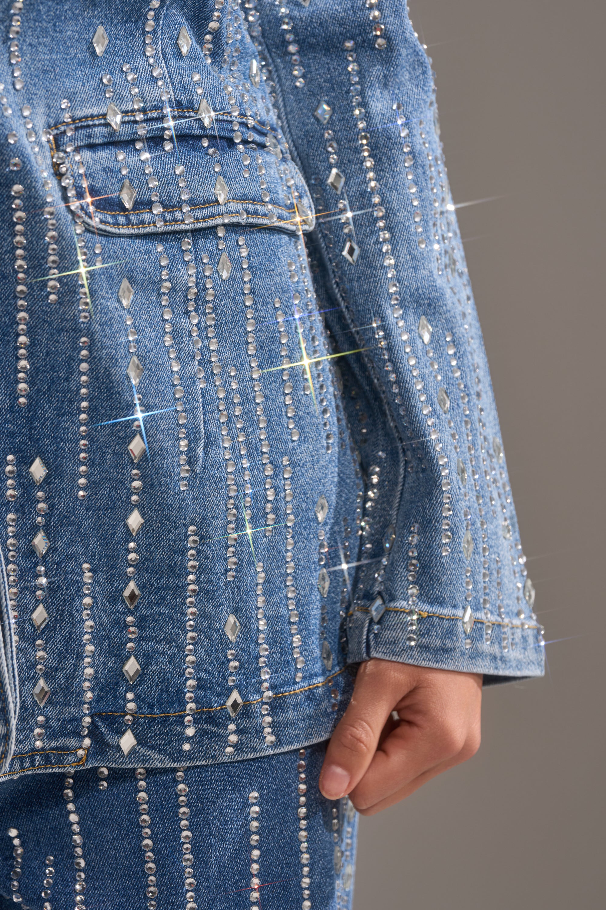 Close-up of a person wearing the NEVER COME DOWN RHINESTONE DENIM BLAZER and matching jeans, featuring vertical rows of sparkling rhinestones, with one hand at their side against a neutral background.