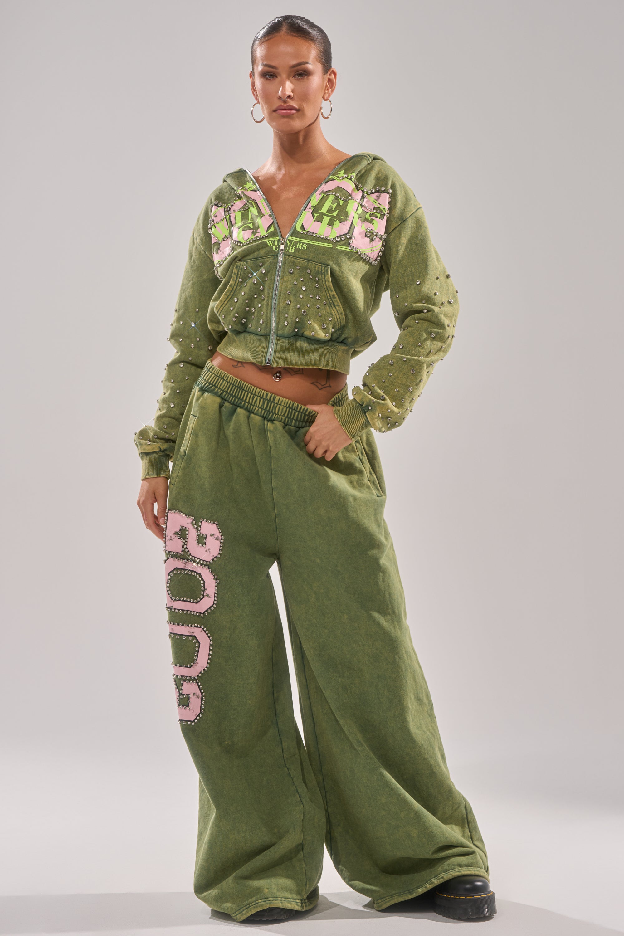 A woman stands confidently in Y2K-inspired loungewear: the WINNERS CLUB WIDE LEG JOGGER, an olive green matching set with pink and white "2000" patches, featuring a zip-up jacket and wide leg joggers. 0