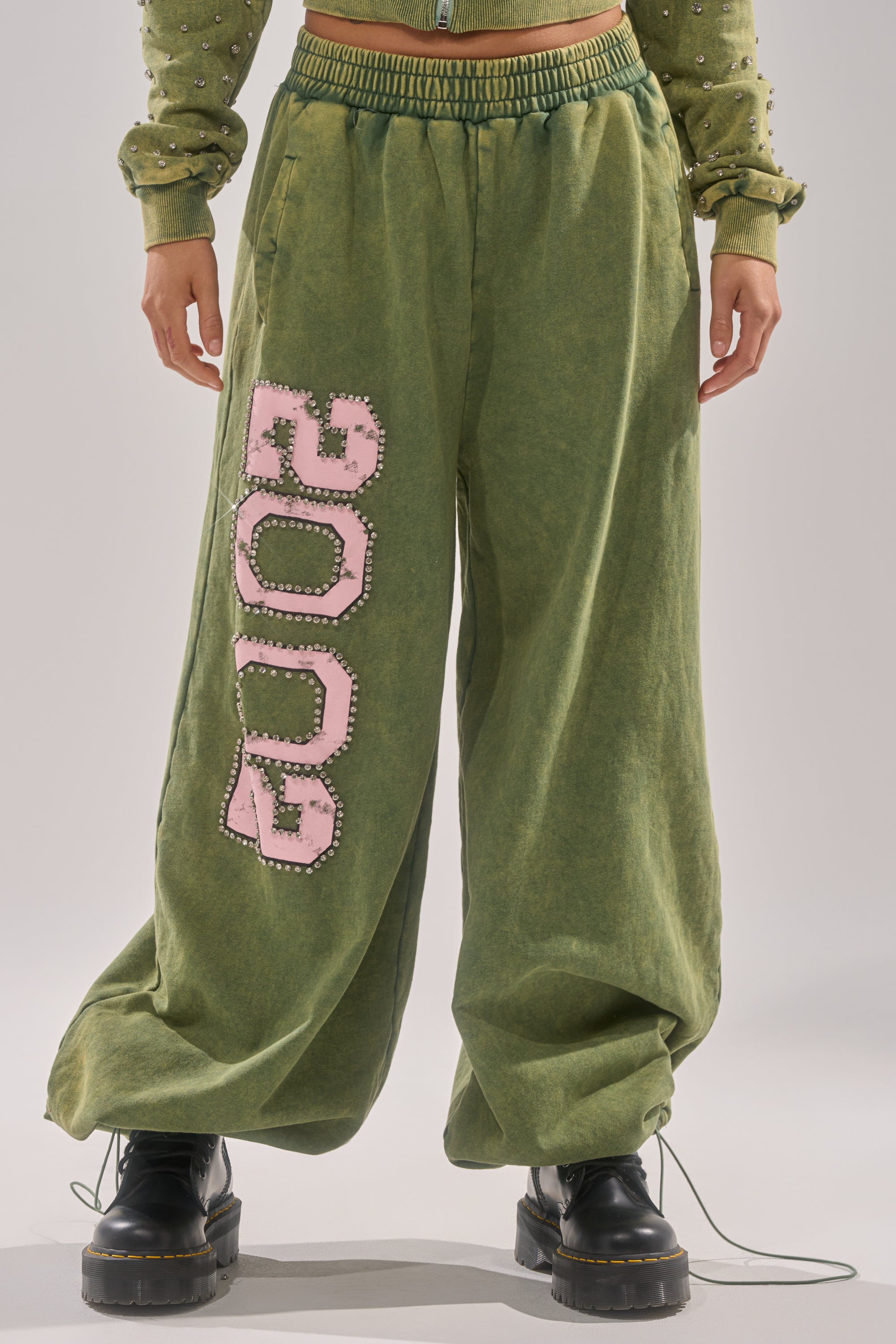 A person wearing WINNERS CLUB WIDE LEG JOGGER sweatpants in Y2K-inspired green with bold pink “2002,” paired with a matching sweatshirt and black platform boots. Only the lower half of the body is shown. 2
