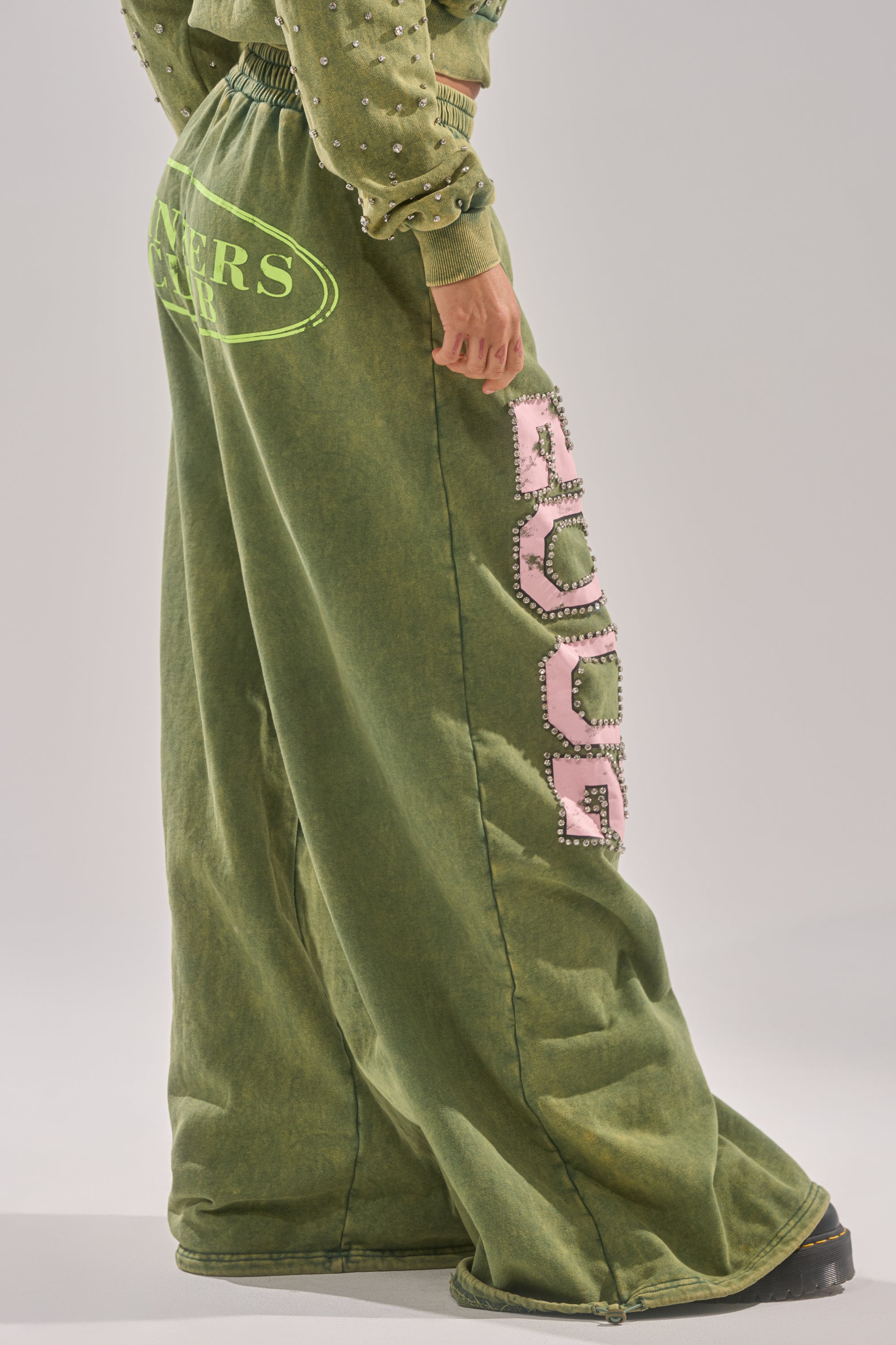 Someone wears the WINNERS CLUB WIDE LEG JOGGER: a Y2K-inspired, loose-fit, faded green jogger with pink studded lettering down the leg and a matching green sweatshirt sleeve. The background is plain and light-colored. 3