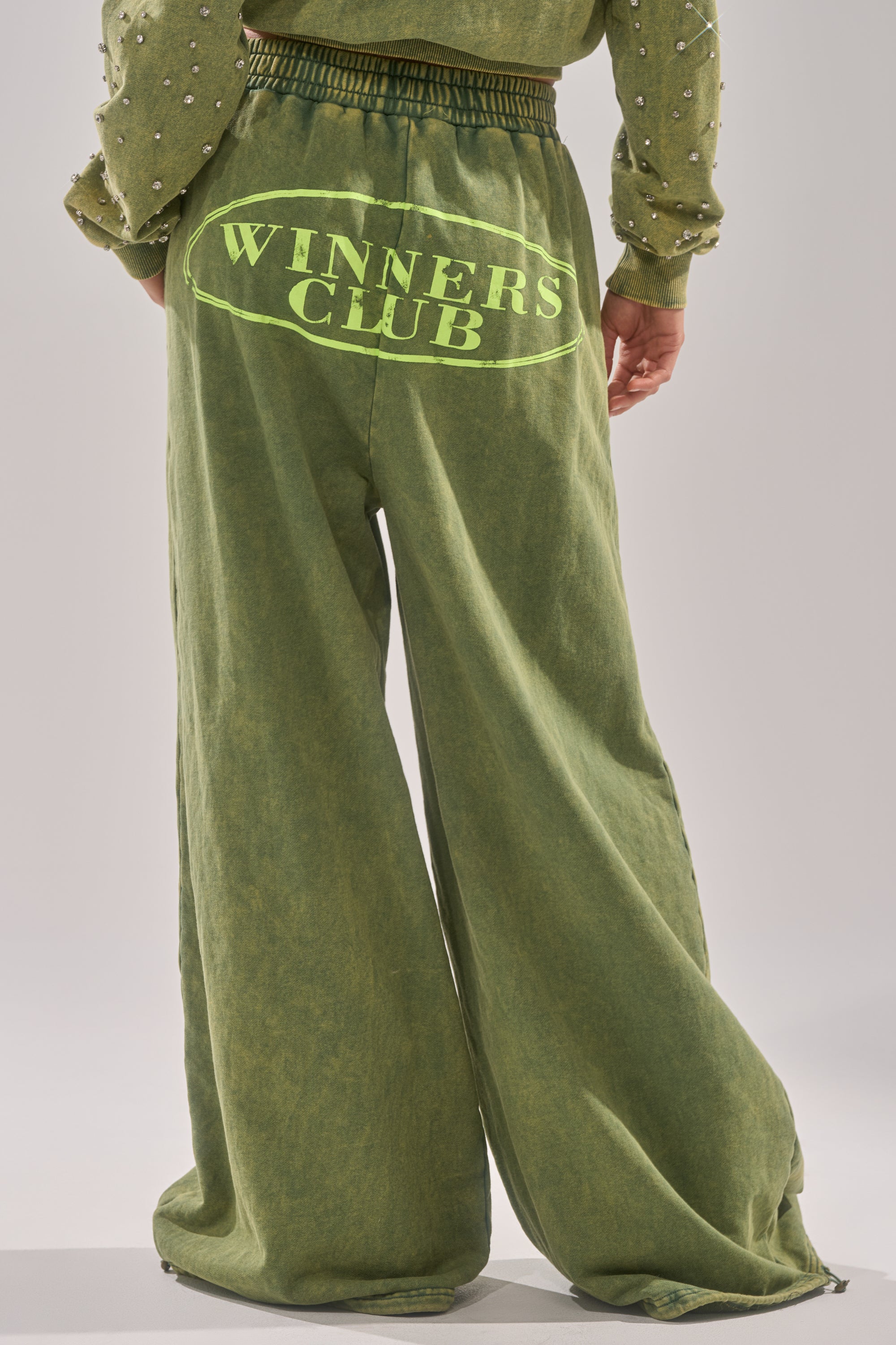 Showcasing only the lower half, a person wears the WINNERS CLUB WIDE LEG JOGGER—Y2K-inspired, loose-fit green joggers with "WINNERS CLUB" in bold yellow on the back—paired with a top featuring pearl-like sleeve details. 4