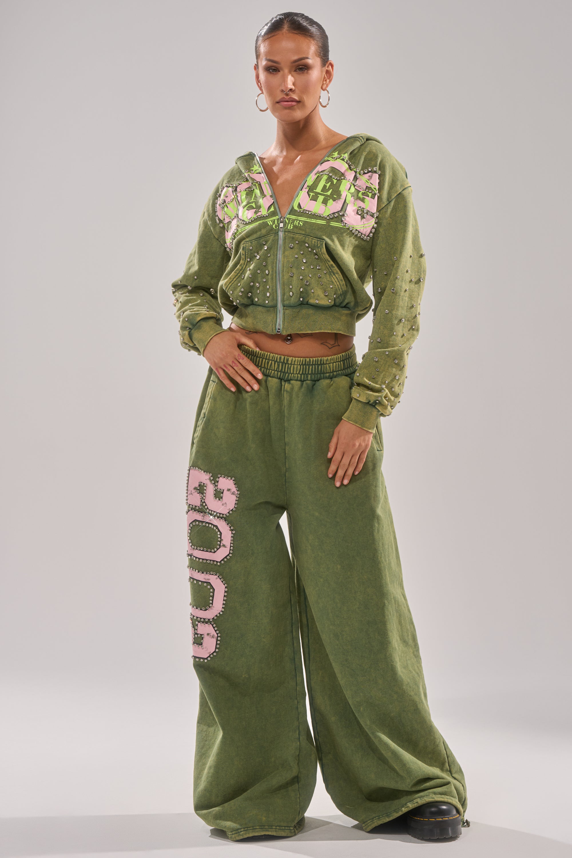 A woman poses confidently in green Y2K loungewear—WINNERS CLUB WIDE LEG JOGGER and a matching zip-up hoodie—featuring pink and white decorative text with rhinestone trim, set against a plain light background. 5