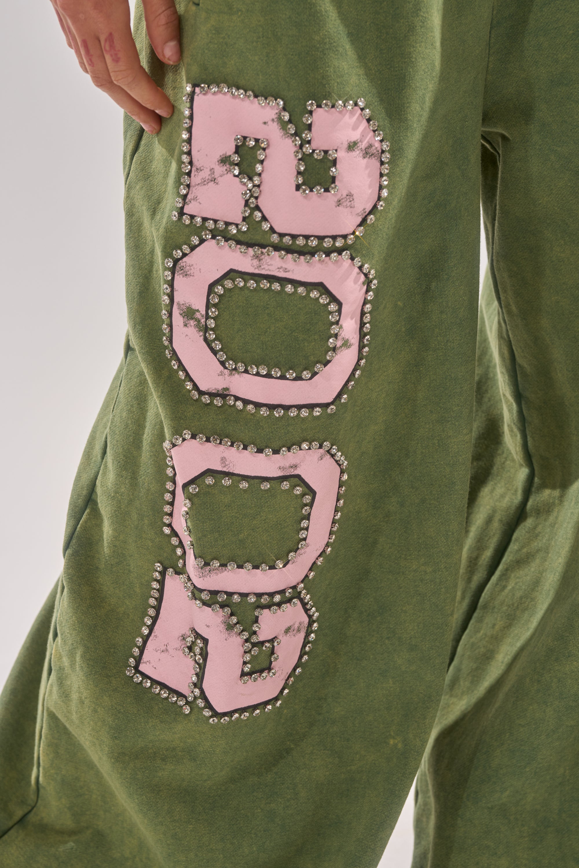 A person wears the WINNERS CLUB WIDE LEG JOGGER, featuring green wide legs with large pink patches outlined in rhinestones that spell "GOOD" vertically. Only part of their hand and the side of their body are visible. 6