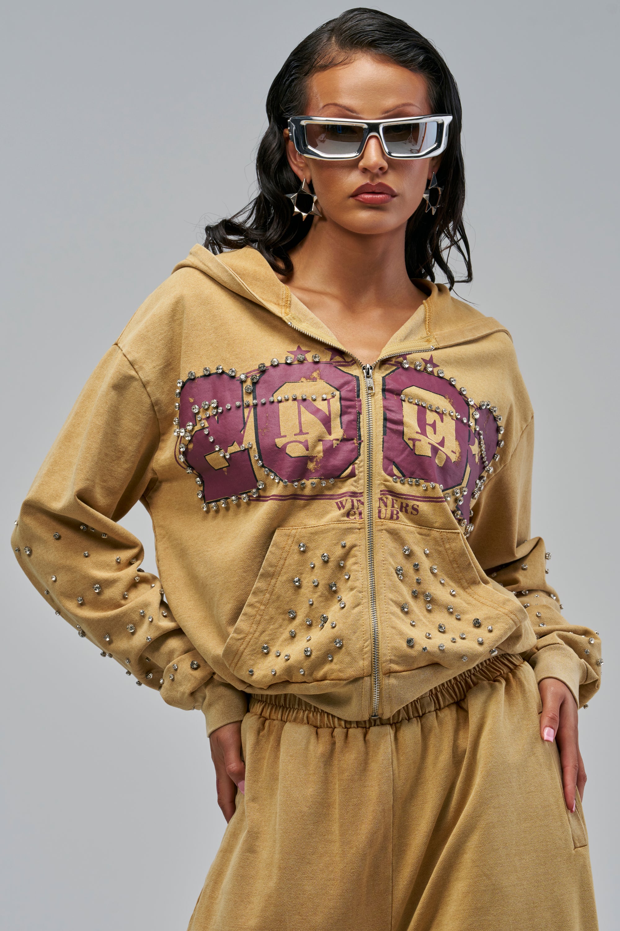 WINNERS CLUB ZIP UP SWEATSHIRT IN MUSTARD, a mustard-colored zip-up hoodie featuring purple collegiate-style lettering on the chest and a studded rhinestone design throughout the sleeves and front pockets.