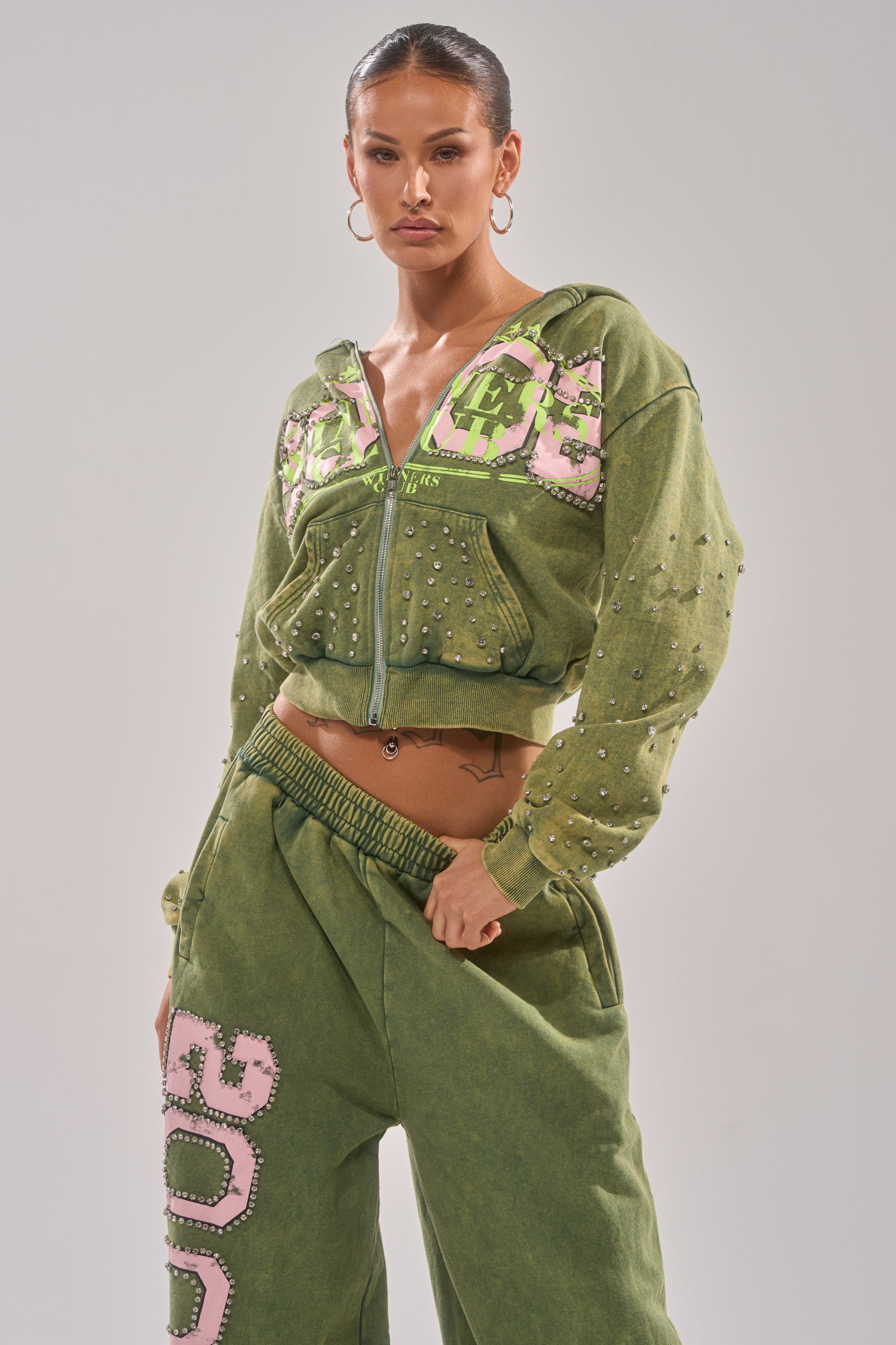 A woman poses confidently in the green WINNERS CLUB ZIP UP SWEATSHIRT and matching pants, featuring pink and white patches, silver studs, rhinestone embellishments, and hoop earrings against a plain light background. 1