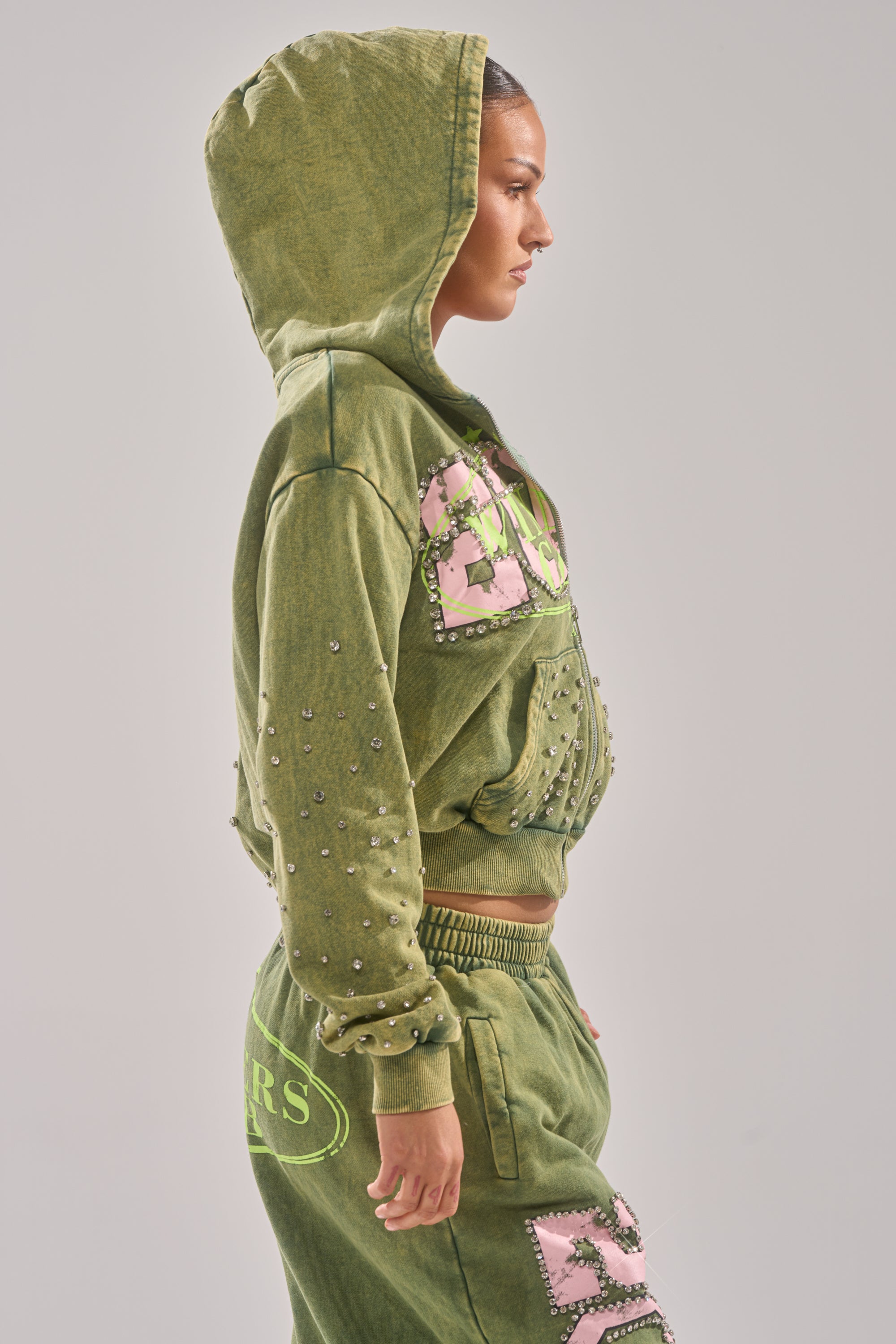 A woman in profile models the WINNERS CLUB ZIP UP SWEATSHIRT—an oversized green hooded sweatshirt with rhinestone and pink patch details—paired with matching sweatpants, set against a plain light gray background. 2