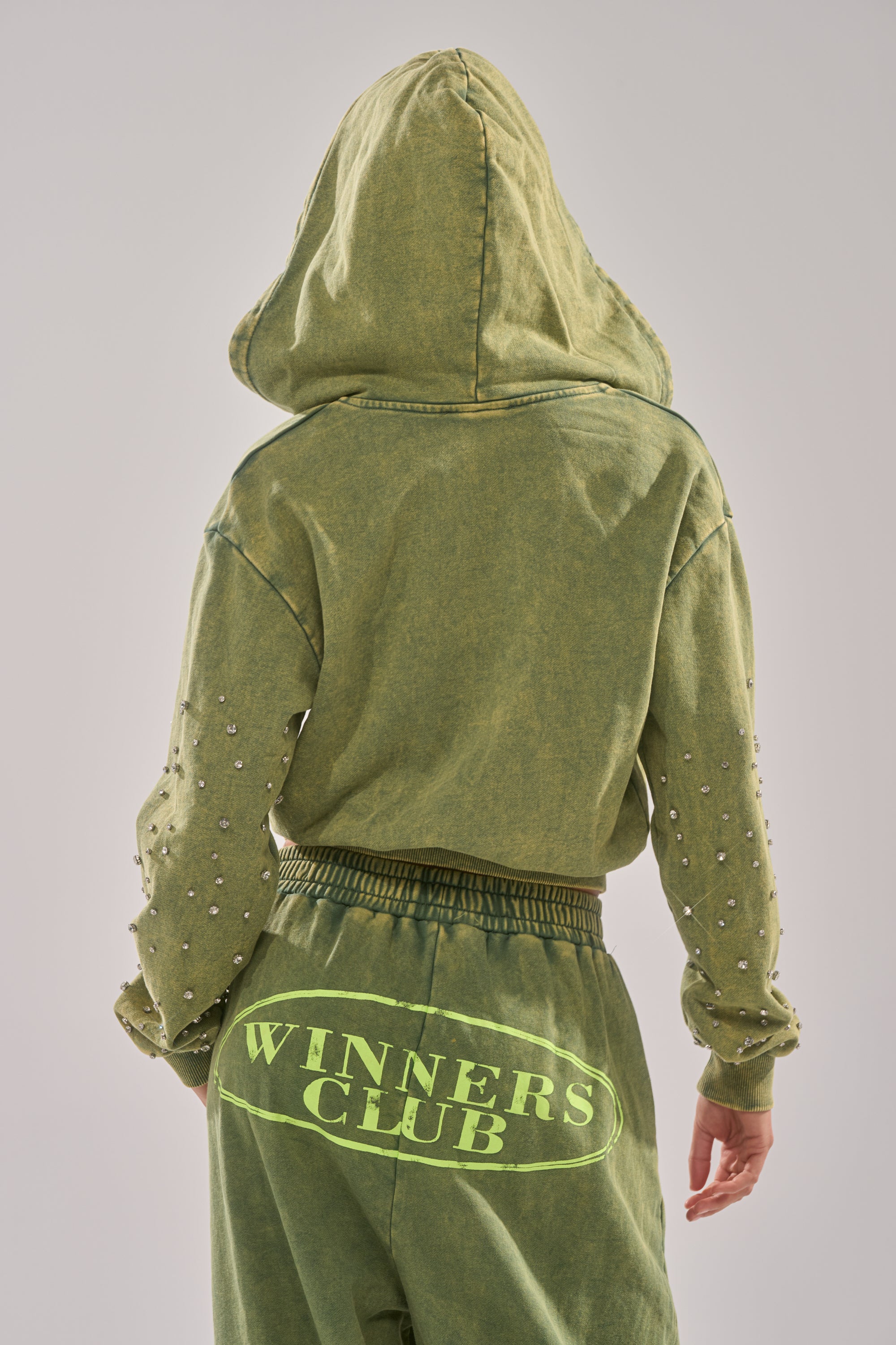 A person faces away wearing an olive green WINNERS CLUB ZIP UP SWEATSHIRT with the hood up and matching joggers. "WINNERS CLUB" is printed large on the back, and the sleeves have small rhinestone embellishments. 3
