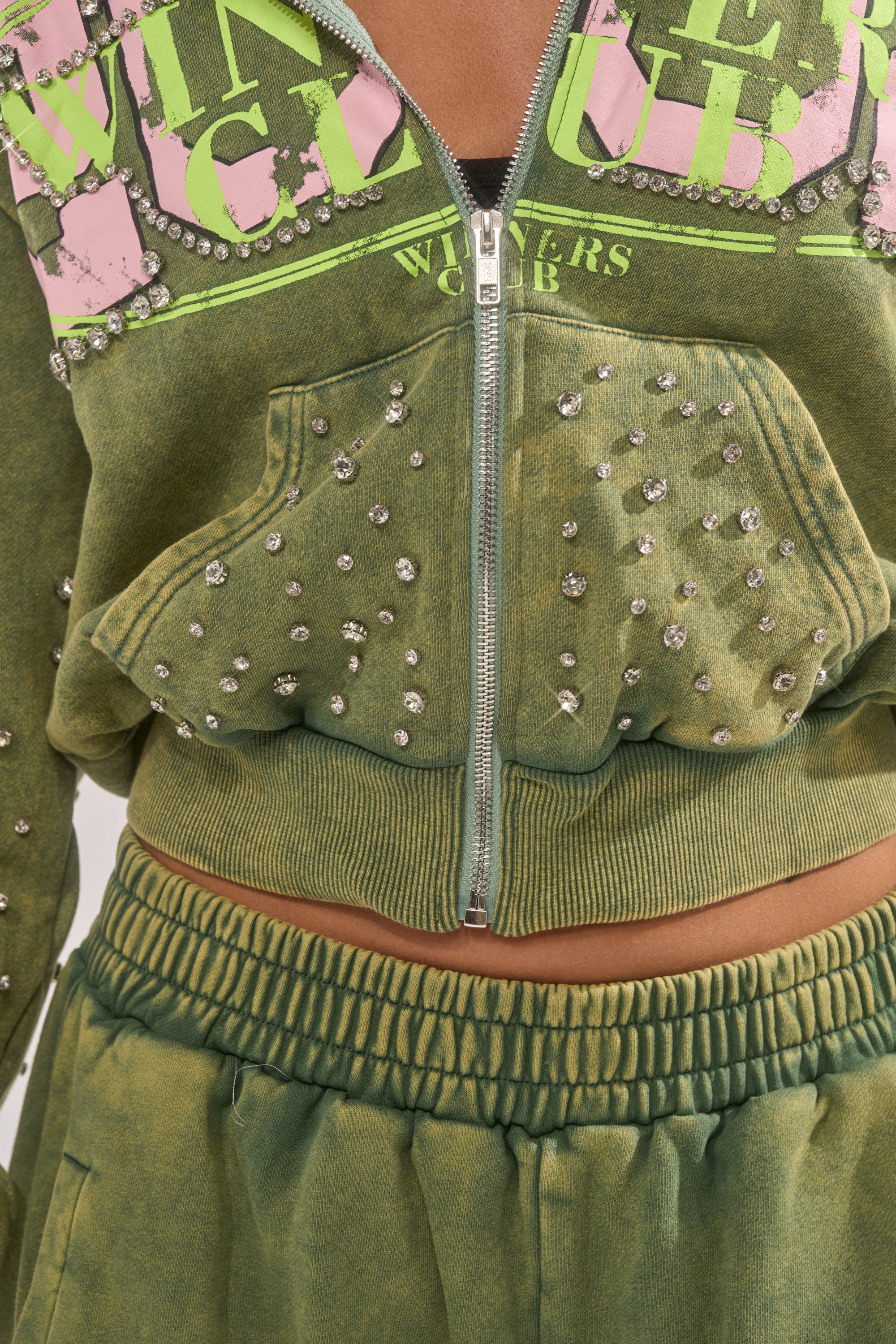 A close-up of a person wearing the WINNERS CLUB ZIP UP SWEATSHIRT, featuring "WINNERS CLUB" text with rhinestone embellishments, studded details, front pocket, and paired with matching green sweatpants. 6