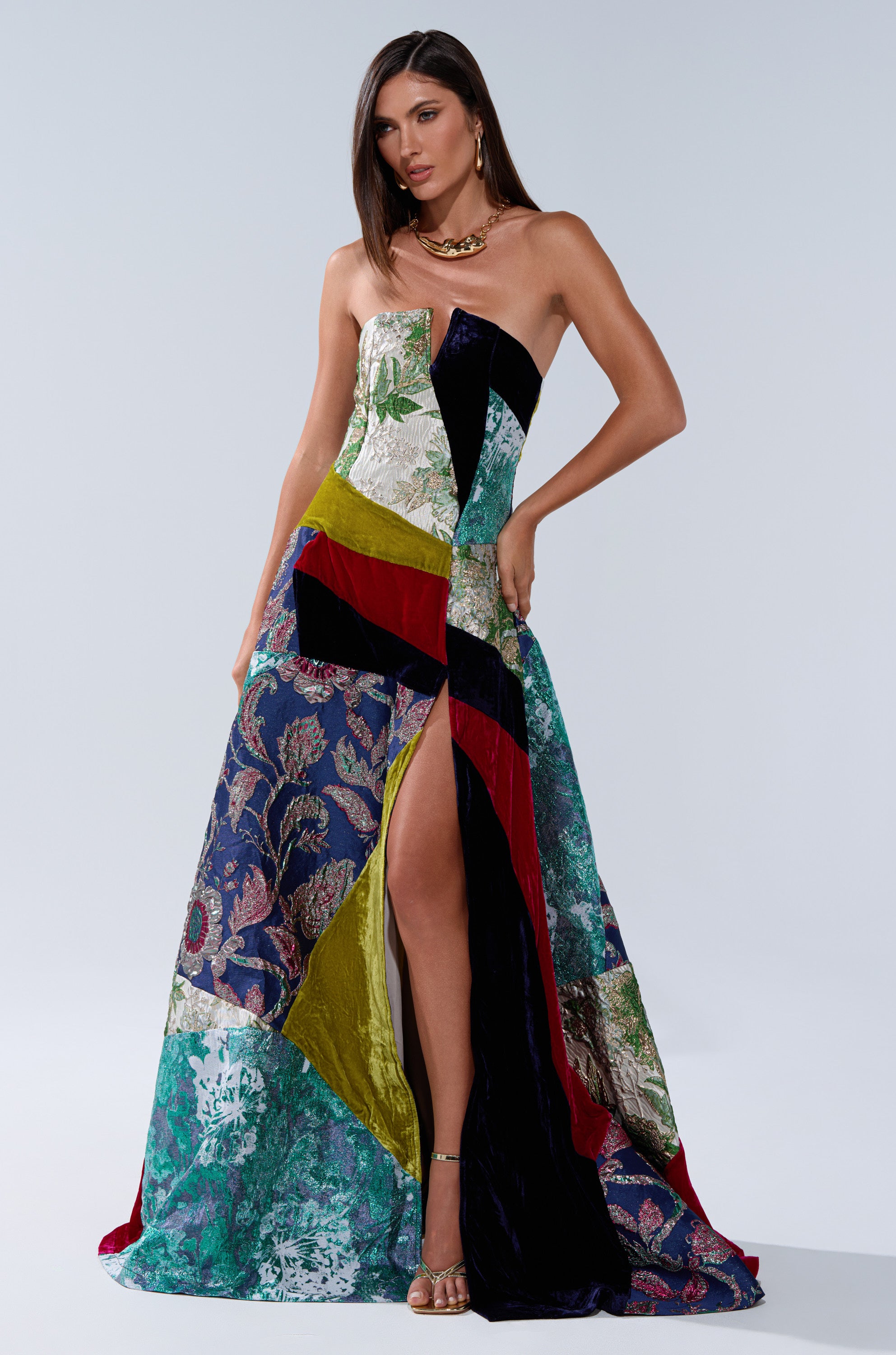 A woman wears the strapless MUSHU MIXED MEDIA MAXI DRESS, a floor-length patchwork gown in jewel-tone green, blue, gold, red, and black velvets. She styles it with gold heels and a chunky gold necklace for an elegant look.