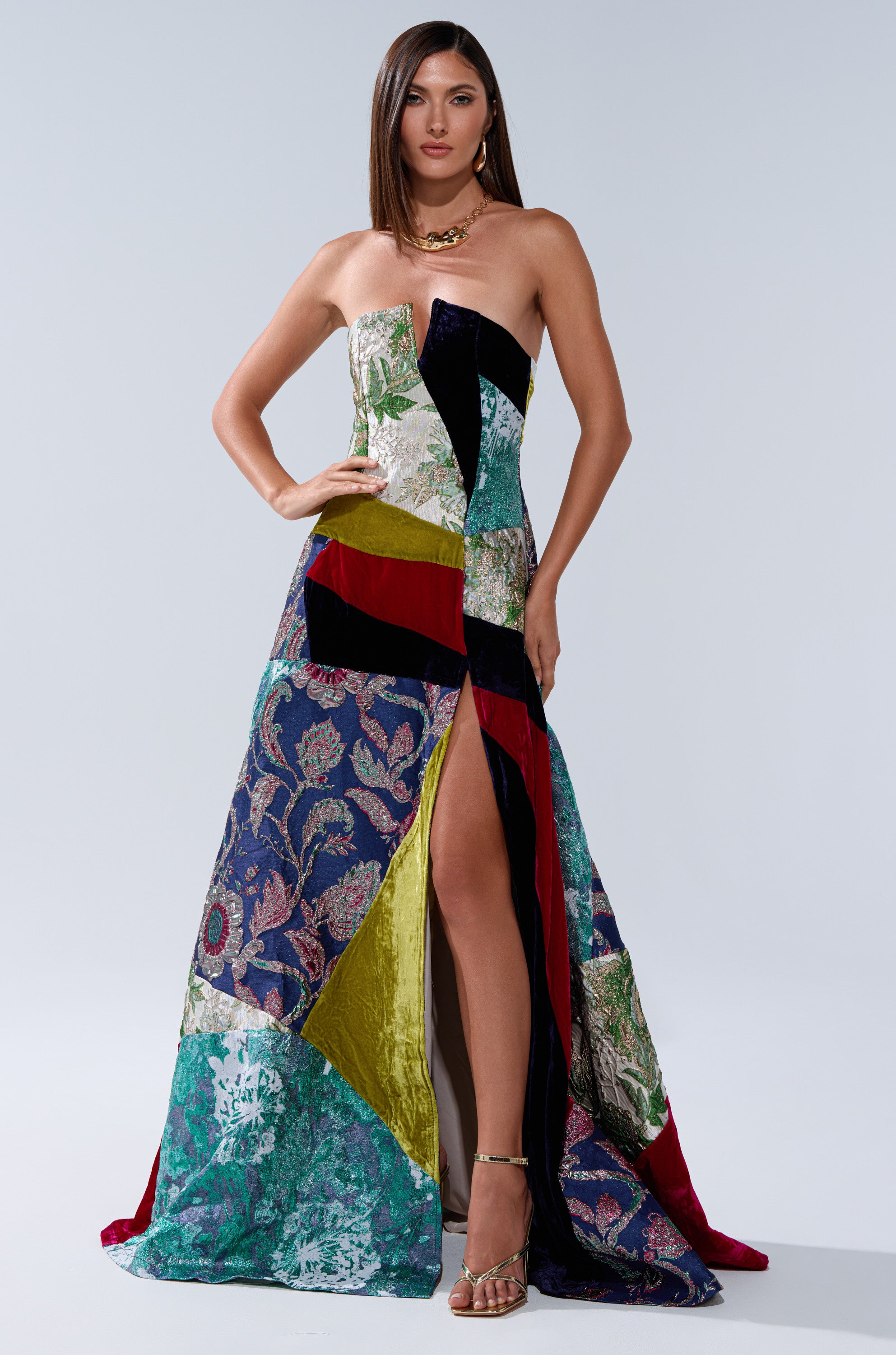A woman exudes confidence in the MUSHU MIXED MEDIA MAXI DRESS, a strapless patchwork maxi with a thigh-high slit. She completes the vibrant look with gold heels, a gold necklace, and sleek straight hair for effortless chic.