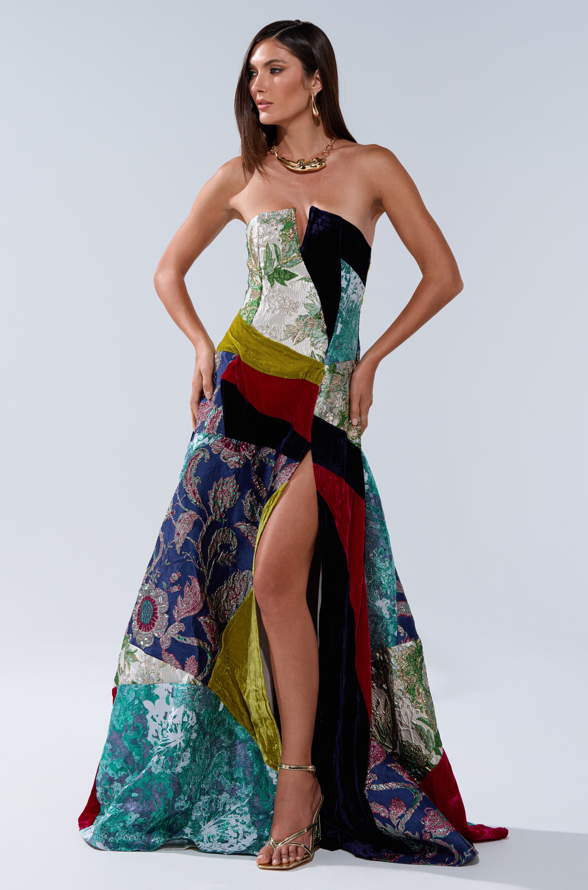 A woman wears the MUSHU MIXED MEDIA MAXI DRESS, a strapless piece with a high slit, showcasing vibrant patchwork—floral, velvet, and geometric patterns. She pairs it with sandals and a bold gold necklace for a standout look.