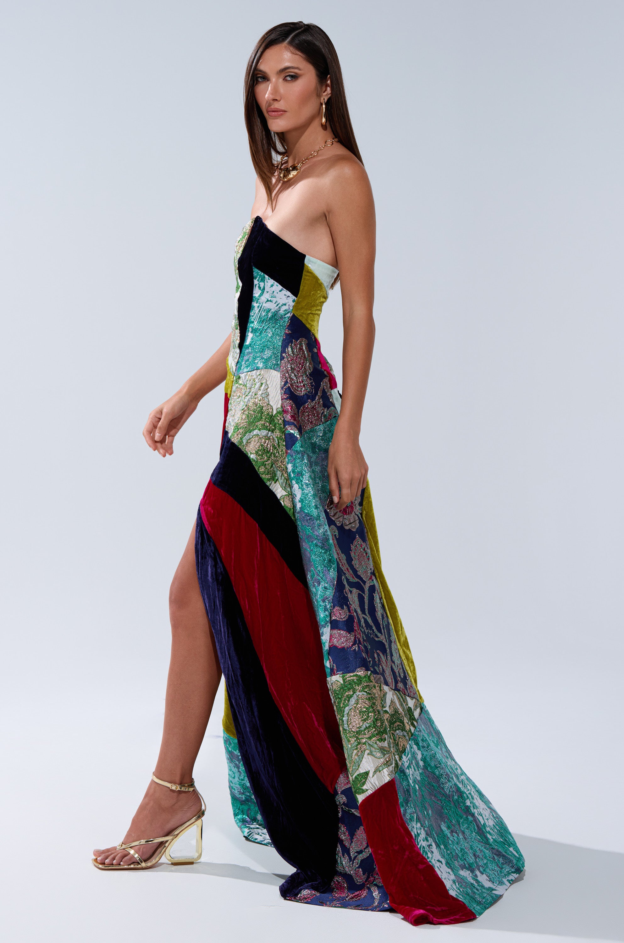 A woman in the MUSHU MIXED MEDIA MAXI DRESS, featuring a strapless design and thigh-high slit, stands in profile. She pairs it with gold strappy heels and jewelry, her straight brown hair framing her face against a plain light backdrop.