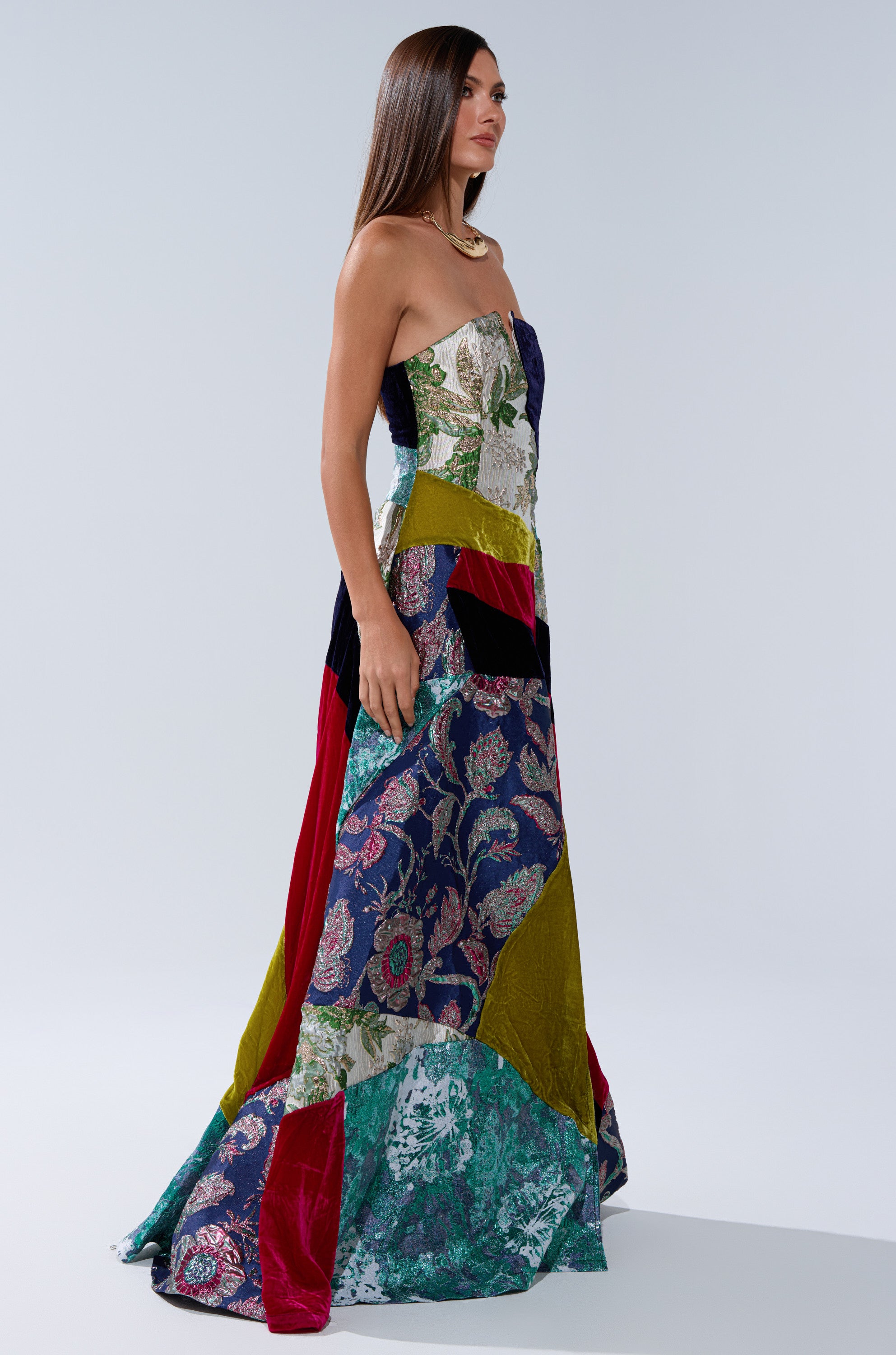 A woman with straight brown hair and a chunky gold necklace stands sideways against a plain light background, wearing the vibrant, avant-garde MUSHU MIXED MEDIA MAXI DRESS featuring floral and velvet patchwork.