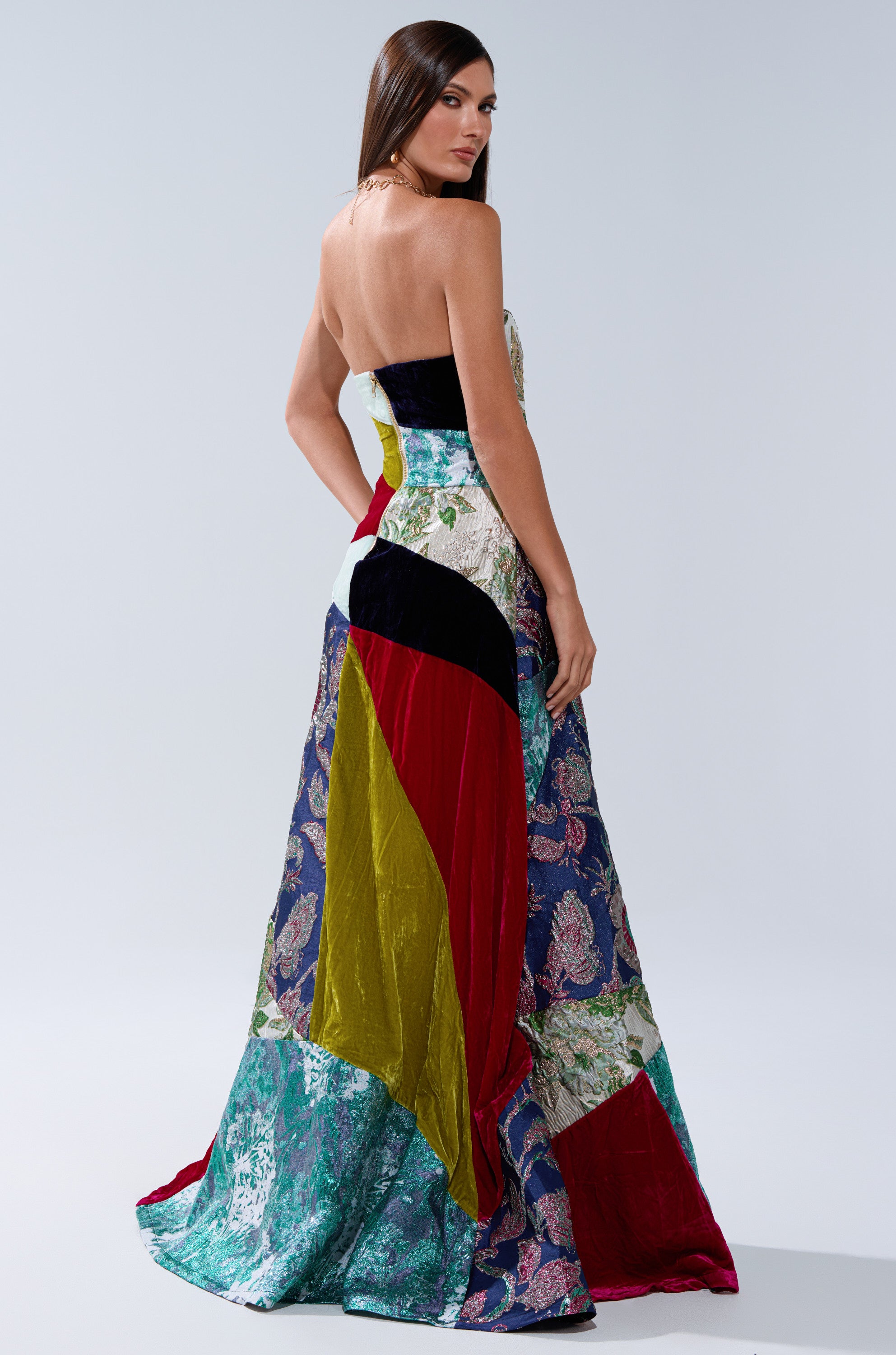 A woman with long brown hair wears the MUSHU MIXED MEDIA MAXI DRESS, a strapless, floor-length piece featuring vibrant green, blue, yellow, red and floral patterns, standing against a plain light gray background—a true statement piece.