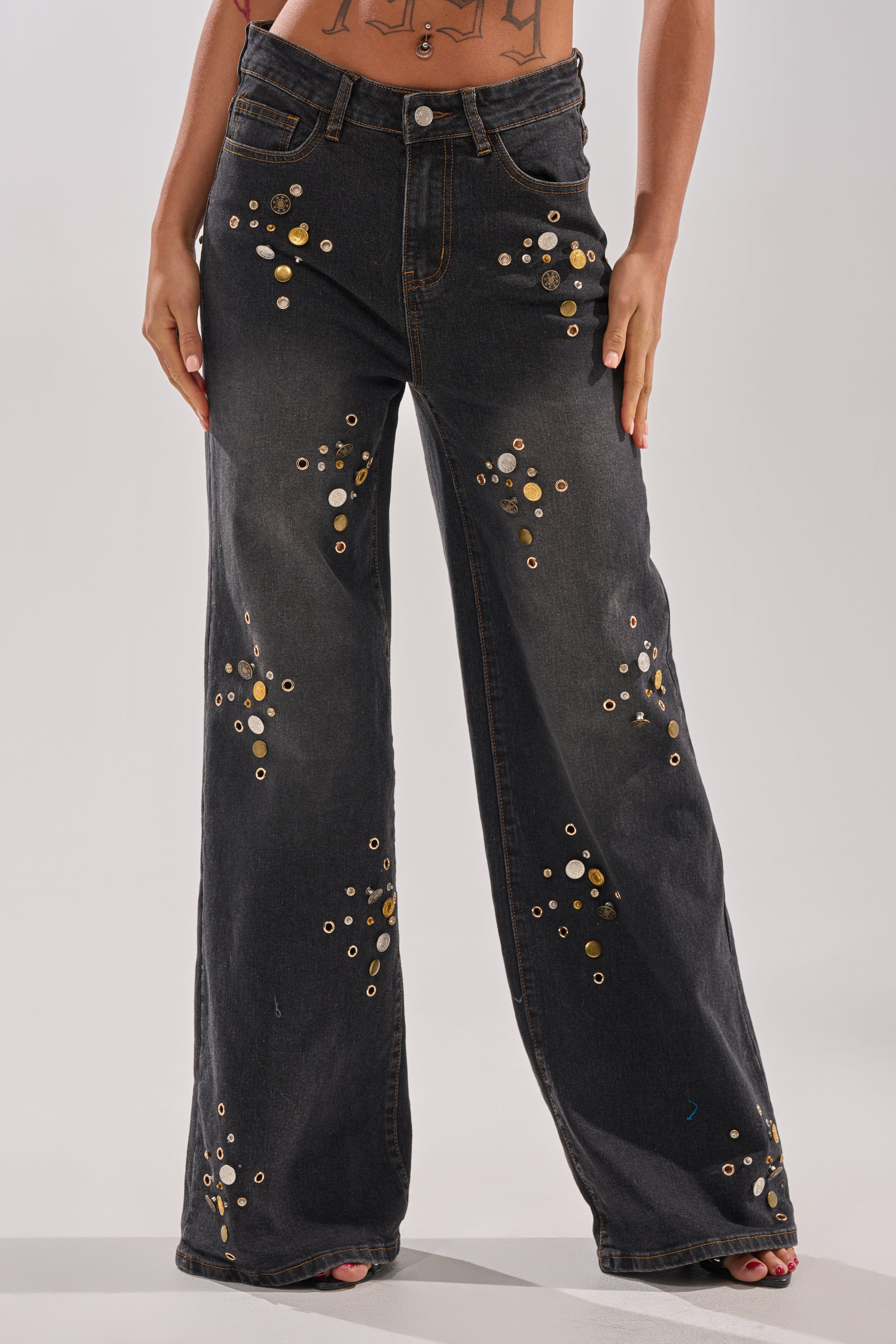 Wearing the SPACE AND TIME STUDDED DENIM—high-rise wide-leg black jeans with gold and silver grommets—a barefoot person stands against a plain, light background, hands on thighs.
