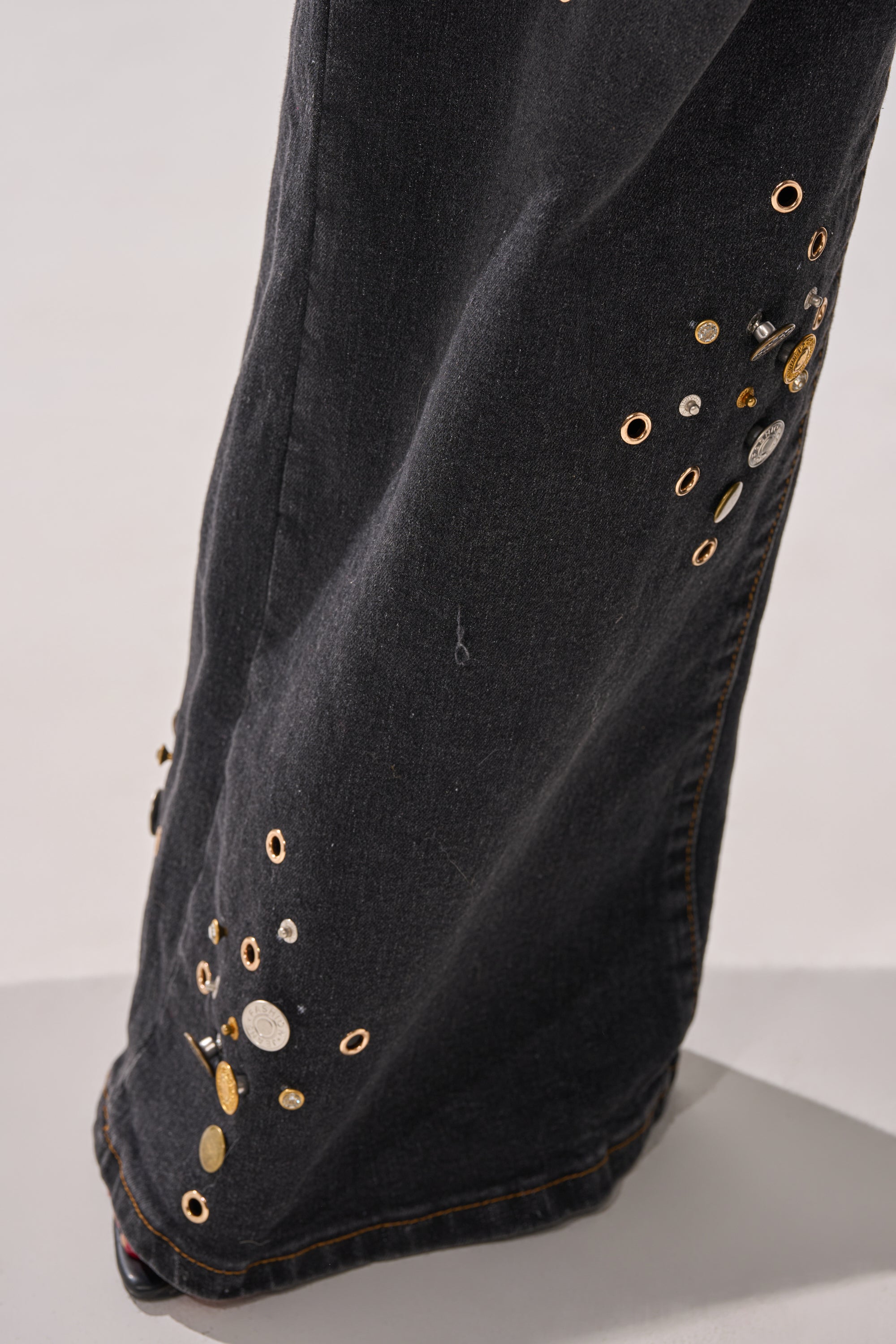 A close-up of the SPACE AND TIME STUDDED DENIM, featuring a high-rise wide-leg jeans silhouette adorned with scattered metallic studs and grommets in gold and silver.
