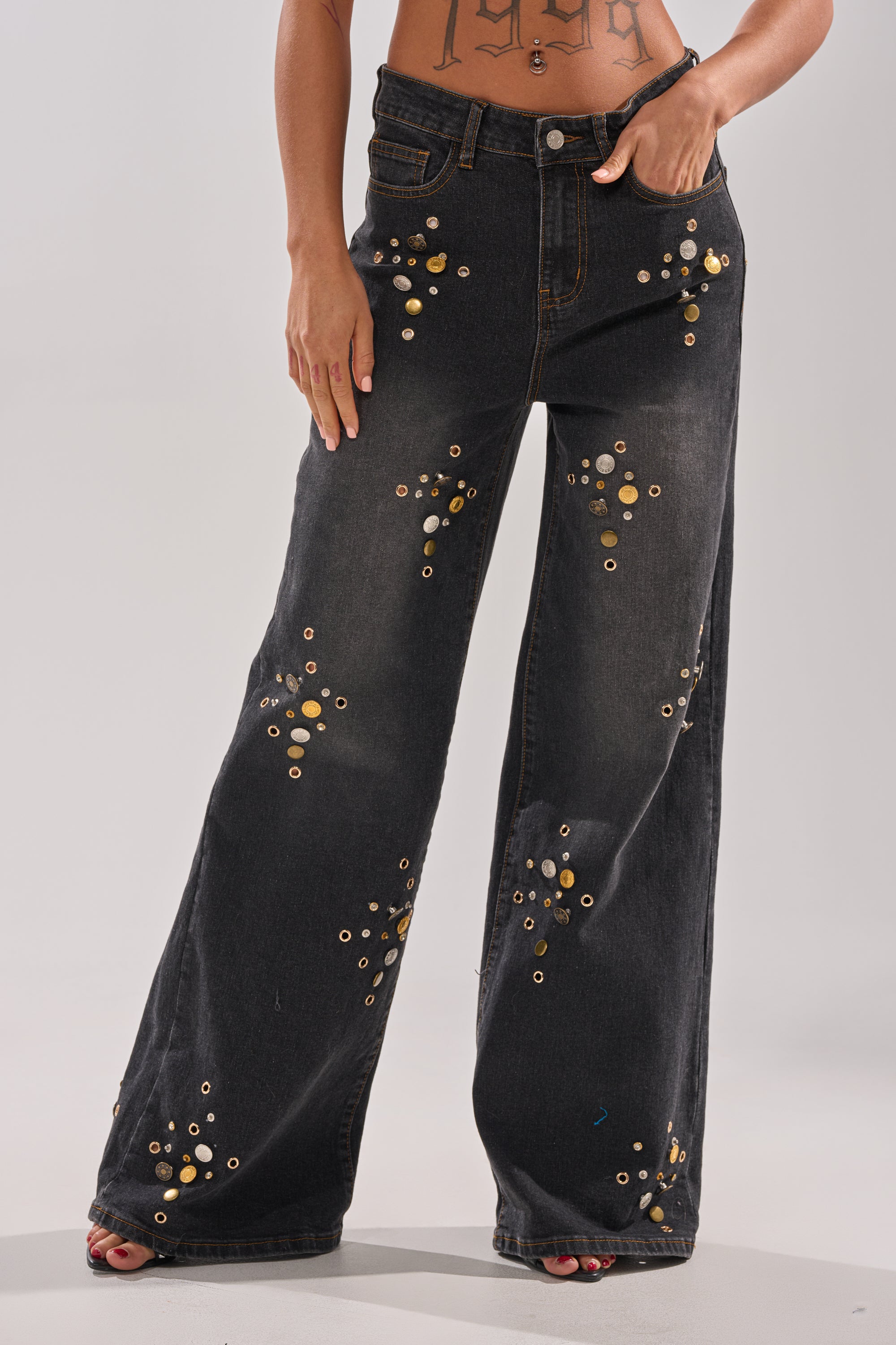 A person models the SPACE AND TIME STUDDED DENIM—high-rise, wide-leg black jeans adorned with gold and silver grommet clusters—standing barefoot with one hand in a pocket and the other on their hip for a bold look.