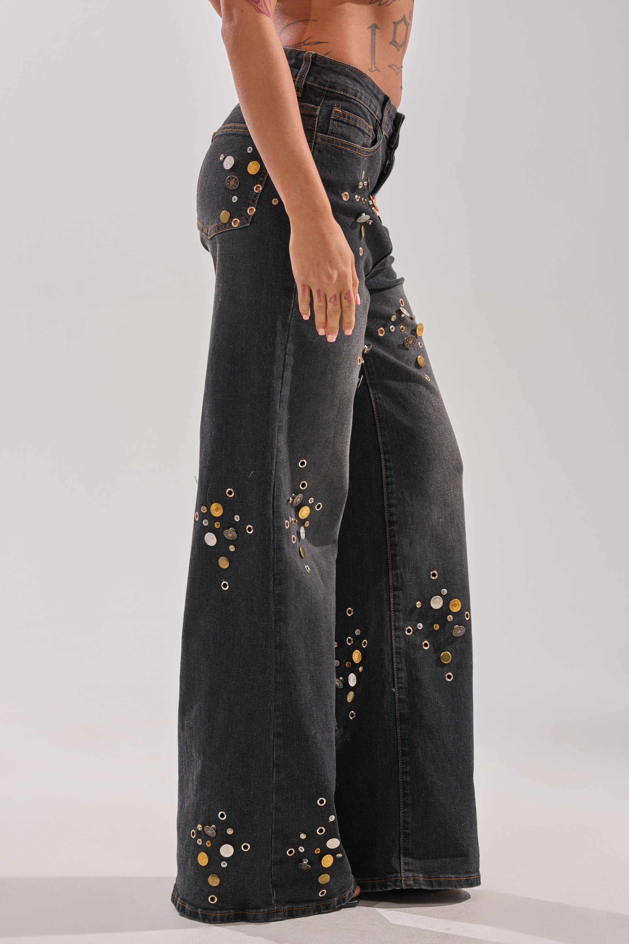 A person models the SPACE AND TIME STUDDED DENIM—black high-rise wide-leg jeans adorned with gold and silver grommets—standing sideways to reveal arm and torso tattoos against a plain light background.
