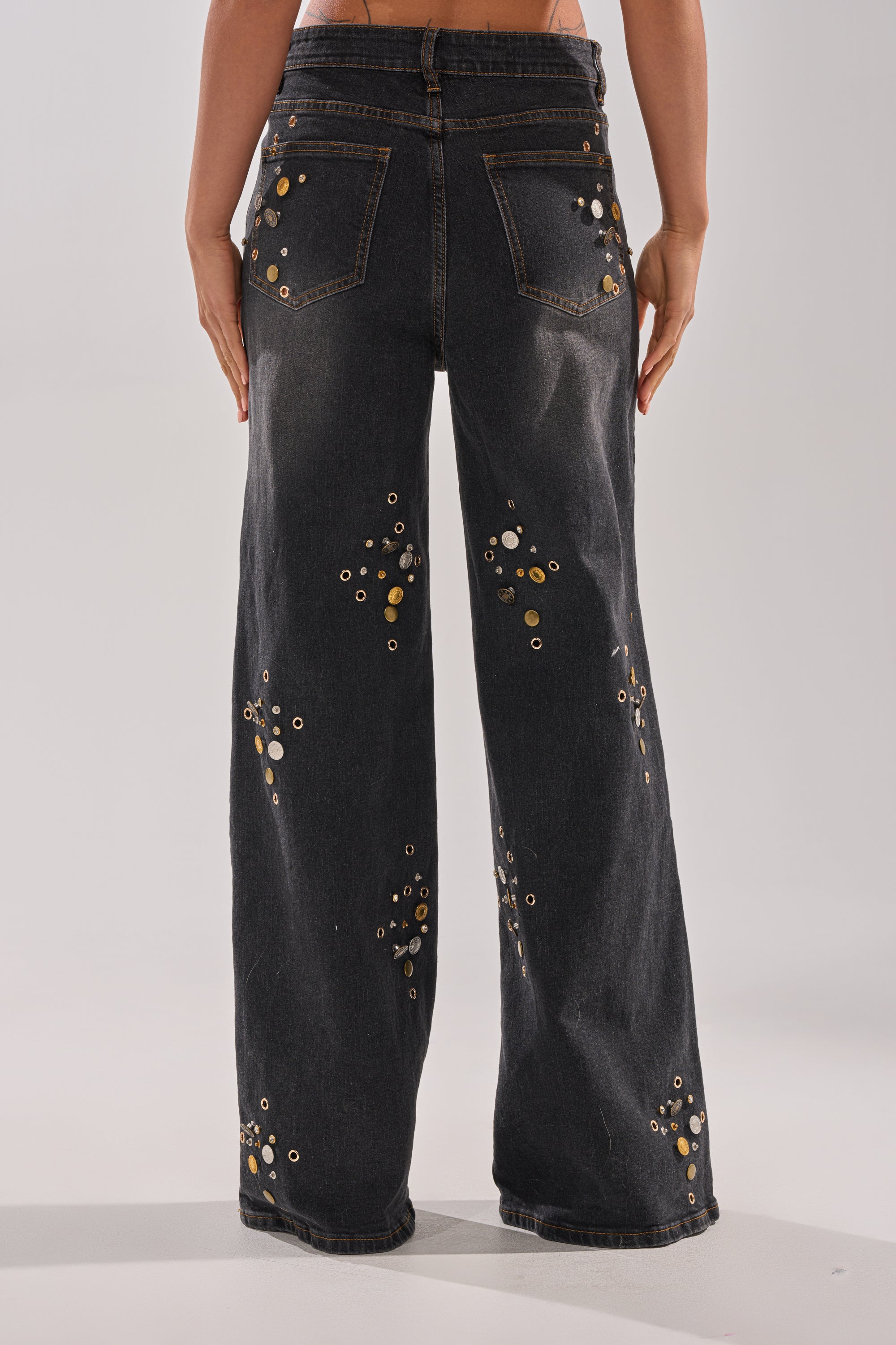 Someone is wearing the SPACE AND TIME STUDDED DENIM, featuring a high-rise fit and wide-leg silhouette with metallic grommet embellishments, photographed from the back against a plain light background.