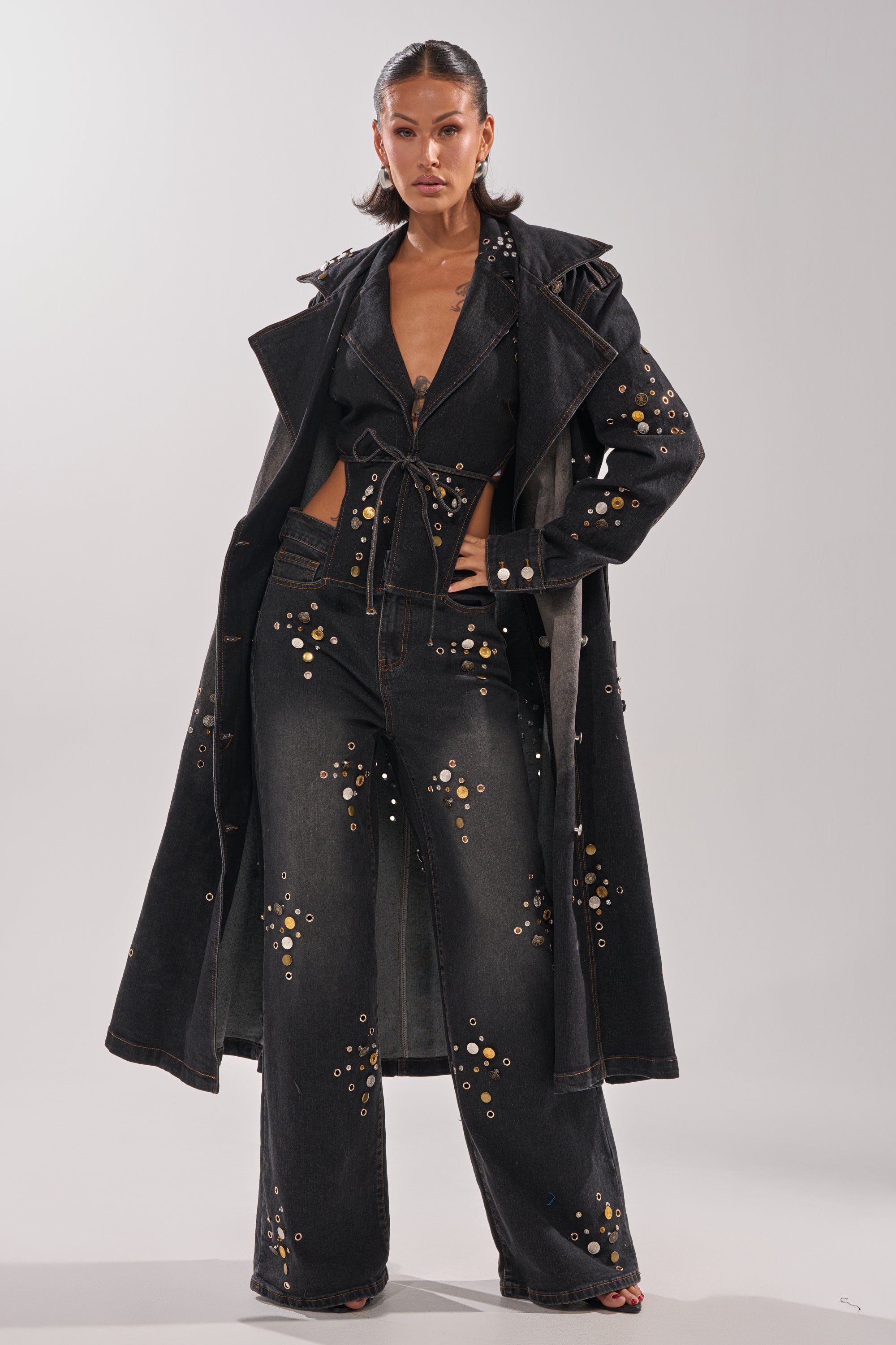 A woman stands confidently in the SPACE AND TIME STUDDED DENIM jumpsuit—high-rise, with gold embellishments—layered under a long black trench coat, posing against a plain light background.