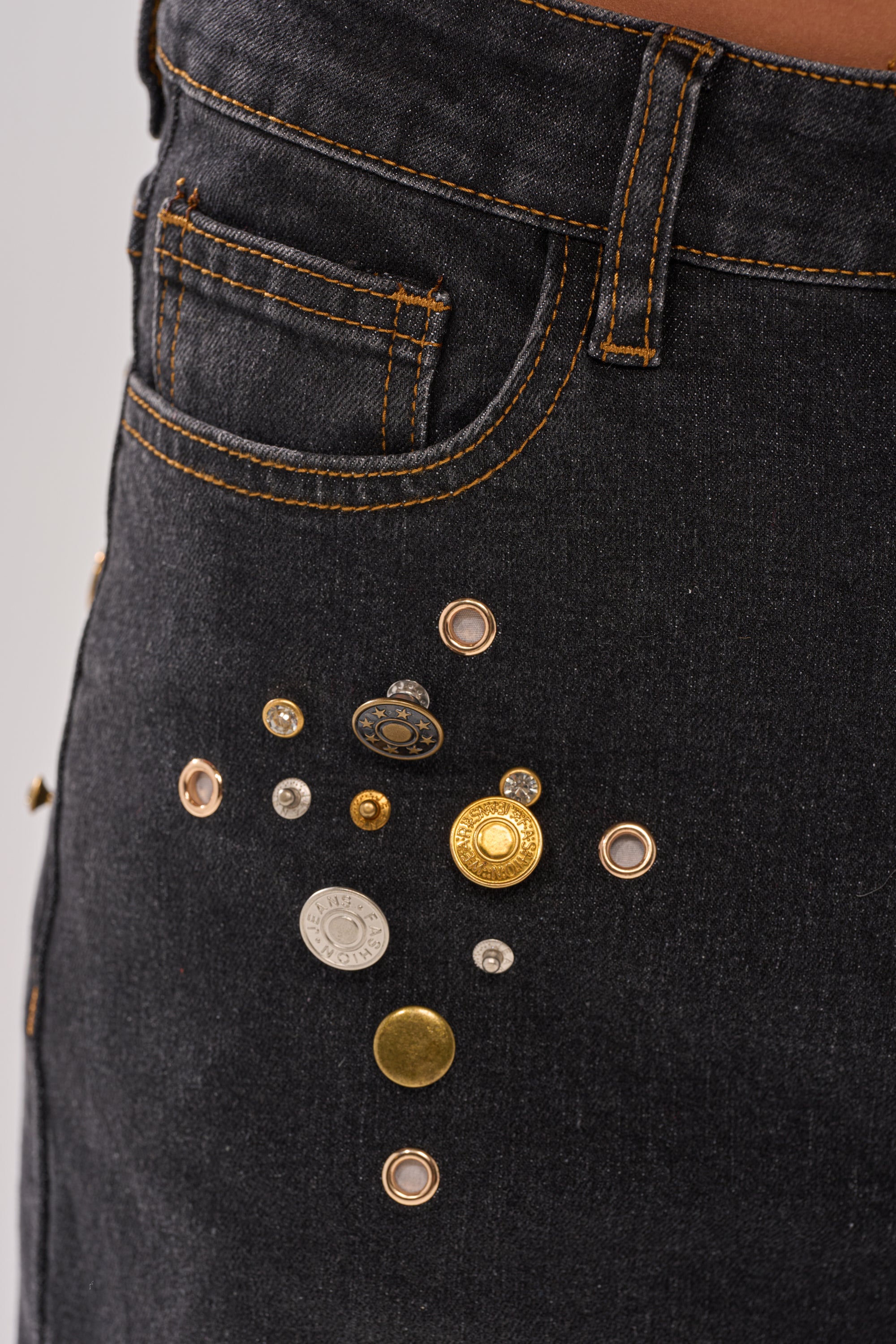 Close-up of SPACE AND TIME STUDDED DENIM: black jeans with orange stitching, adorned with assorted metal buttons and rivets in a plus sign pattern below the front pocket.