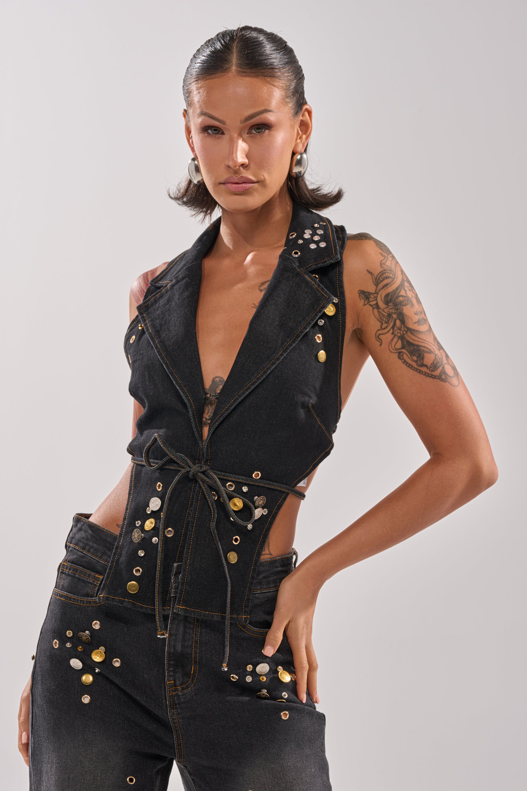 A woman with slicked-back hair and hoop earrings poses confidently in the SPACE AND TIME STUDDED DENIM VEST, a sleeveless dark-wash piece with a deep V-neck, gold studs, visible tattoos, and a chic waist tie.