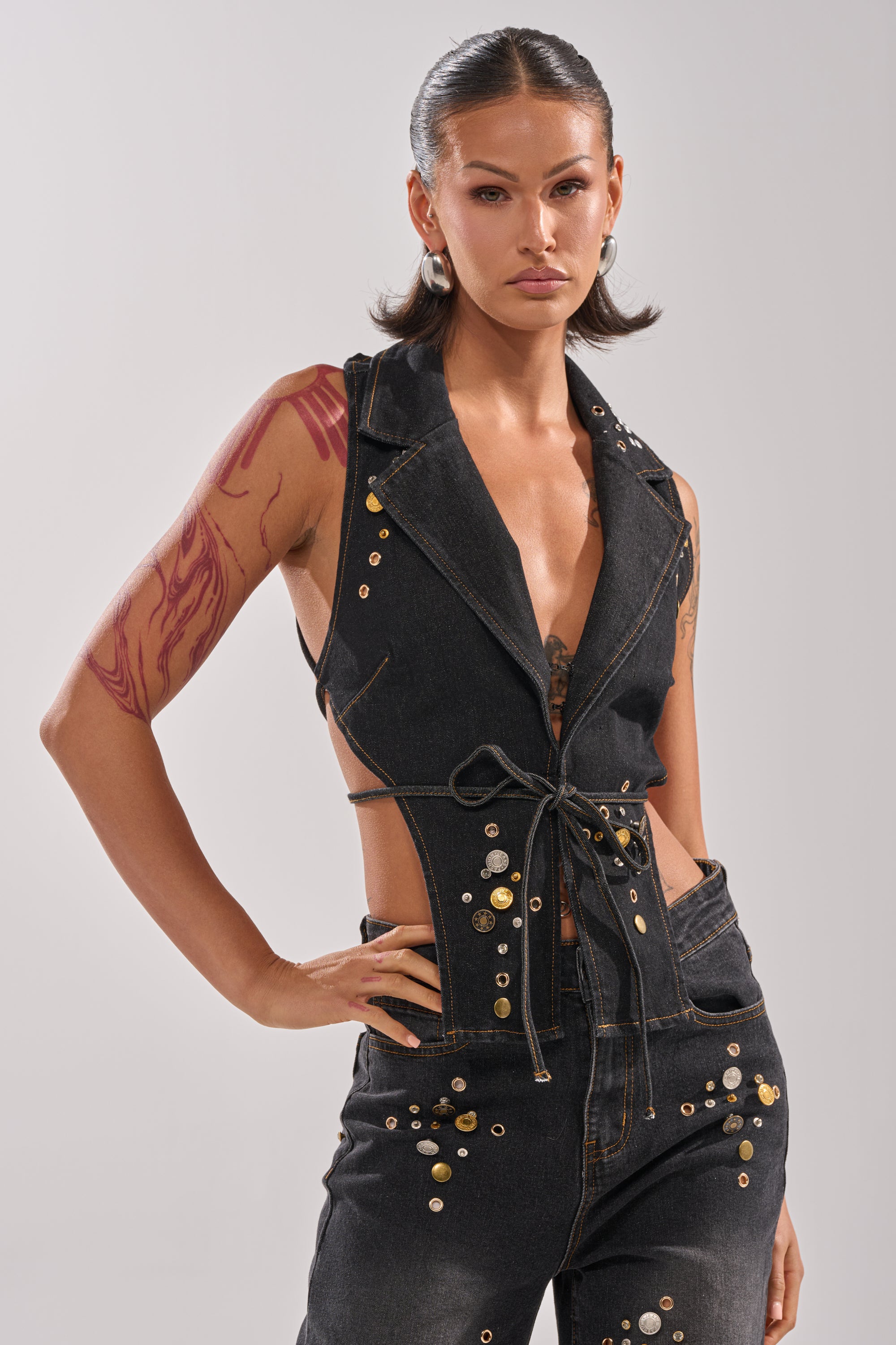 A woman with slicked-back hair models the SPACE AND TIME STUDDED DENIM VEST, featuring a halter neckline, paired with matching high-waisted pants. She displays a red abstract tattoo and poses confidently against a neutral background.