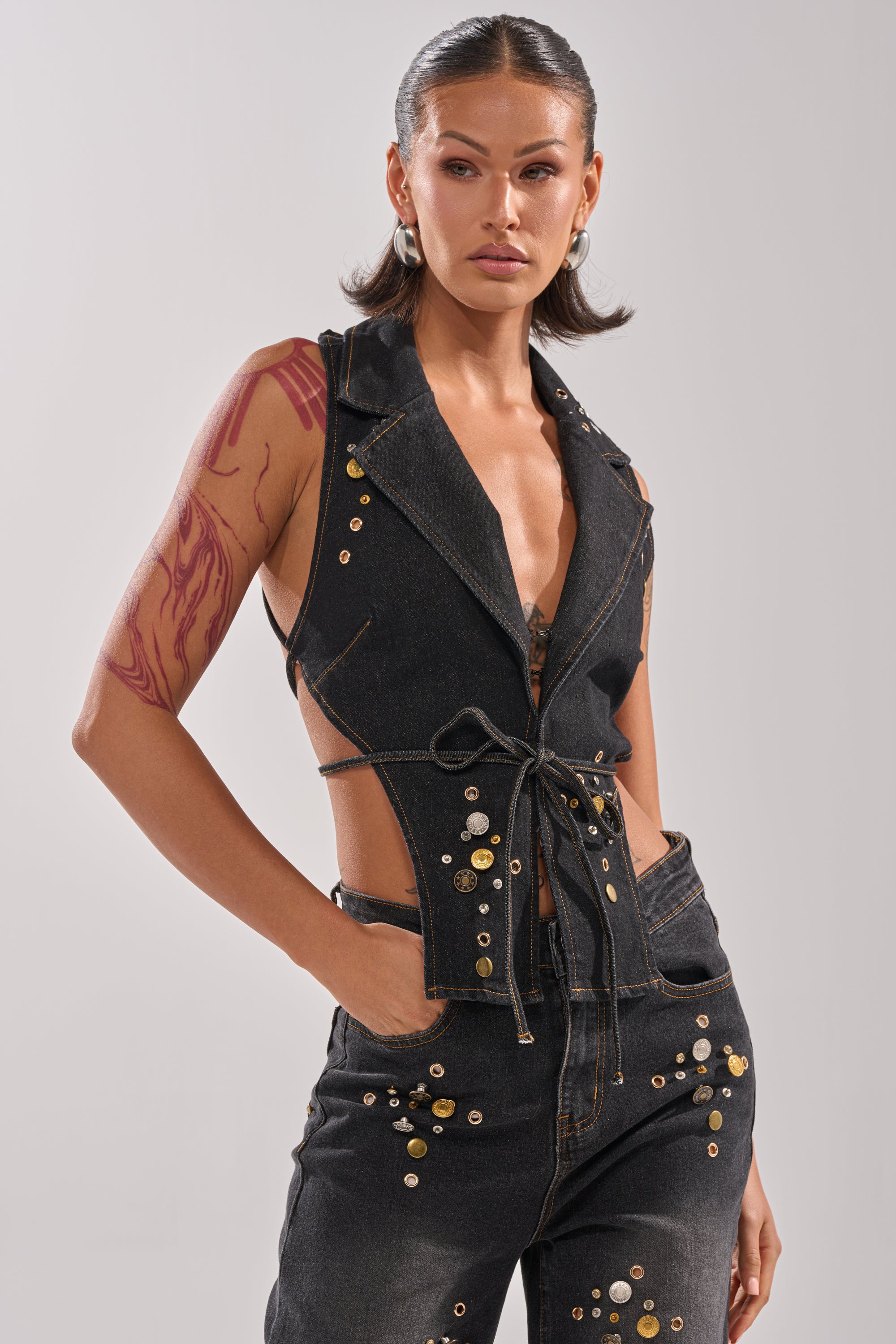 A woman with dark, slicked-back hair rocks the SPACE AND TIME STUDDED DENIM VEST and matching jeans. With one hand in her pocket, a bold red tattoo on her upper arm adds an edgy touch to her confident style.