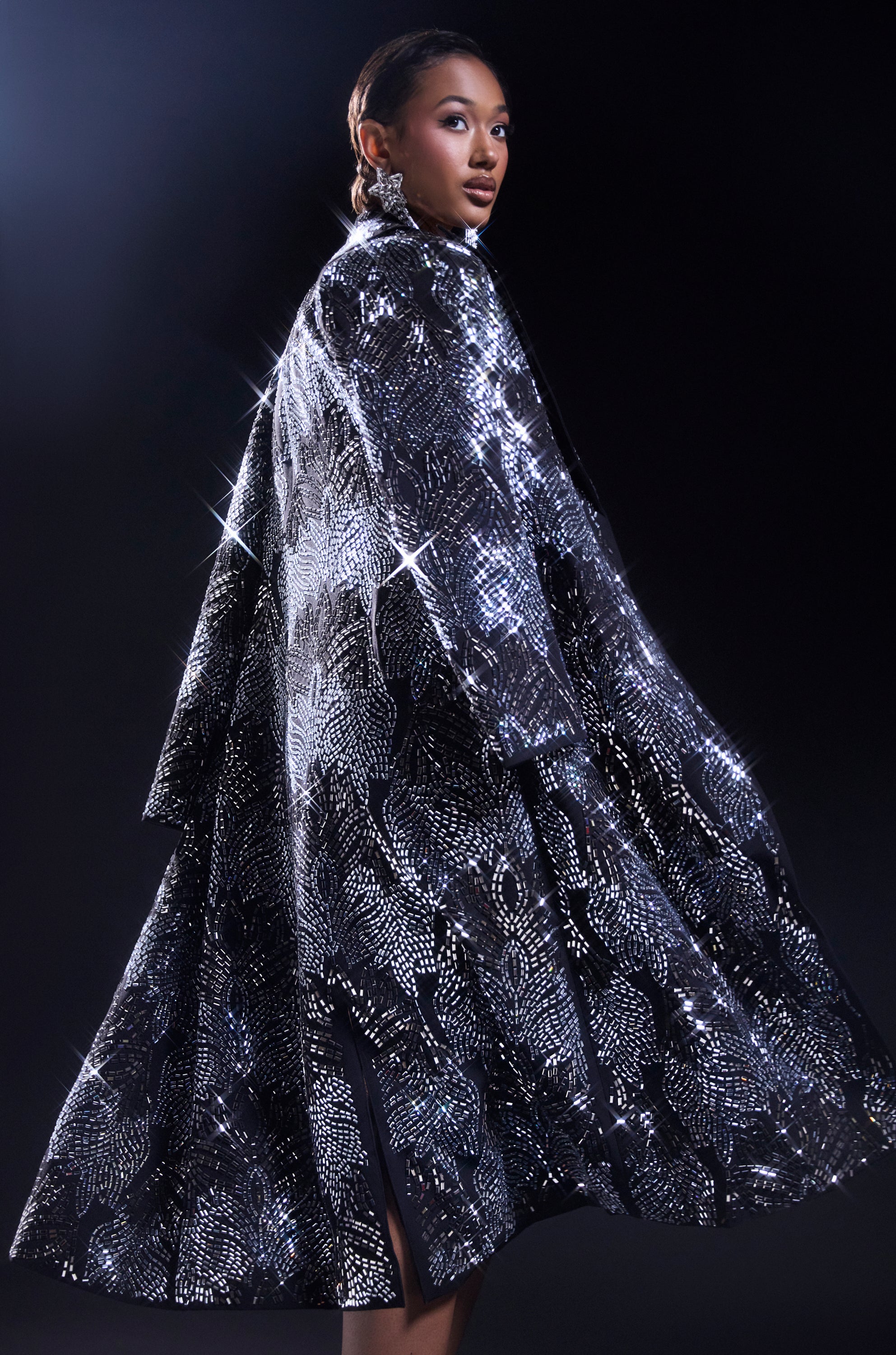A woman poses against a dark background in the SHE IS THE MOMENT RHINESTONE TRENCH, its sparkling embellishments catching the light and creating star-like reflections as she looks over her shoulder. 0