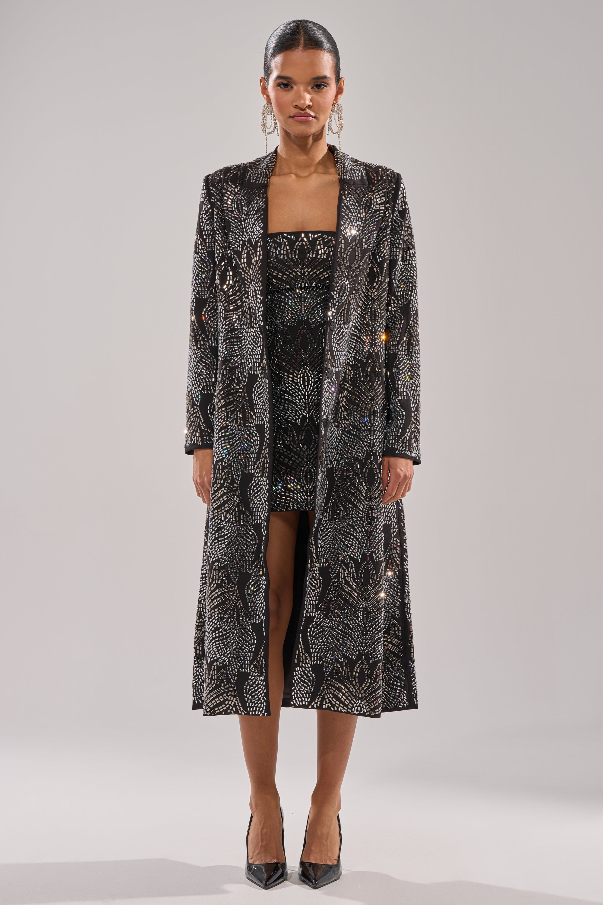 A woman faces forward, wearing a black knee-length dress and the SHE IS THE MOMENT RHINESTONE TRENCH with shimmering silver patterns. She has sleek pulled-back hair, large earrings, and black pointed-toe heels. 4