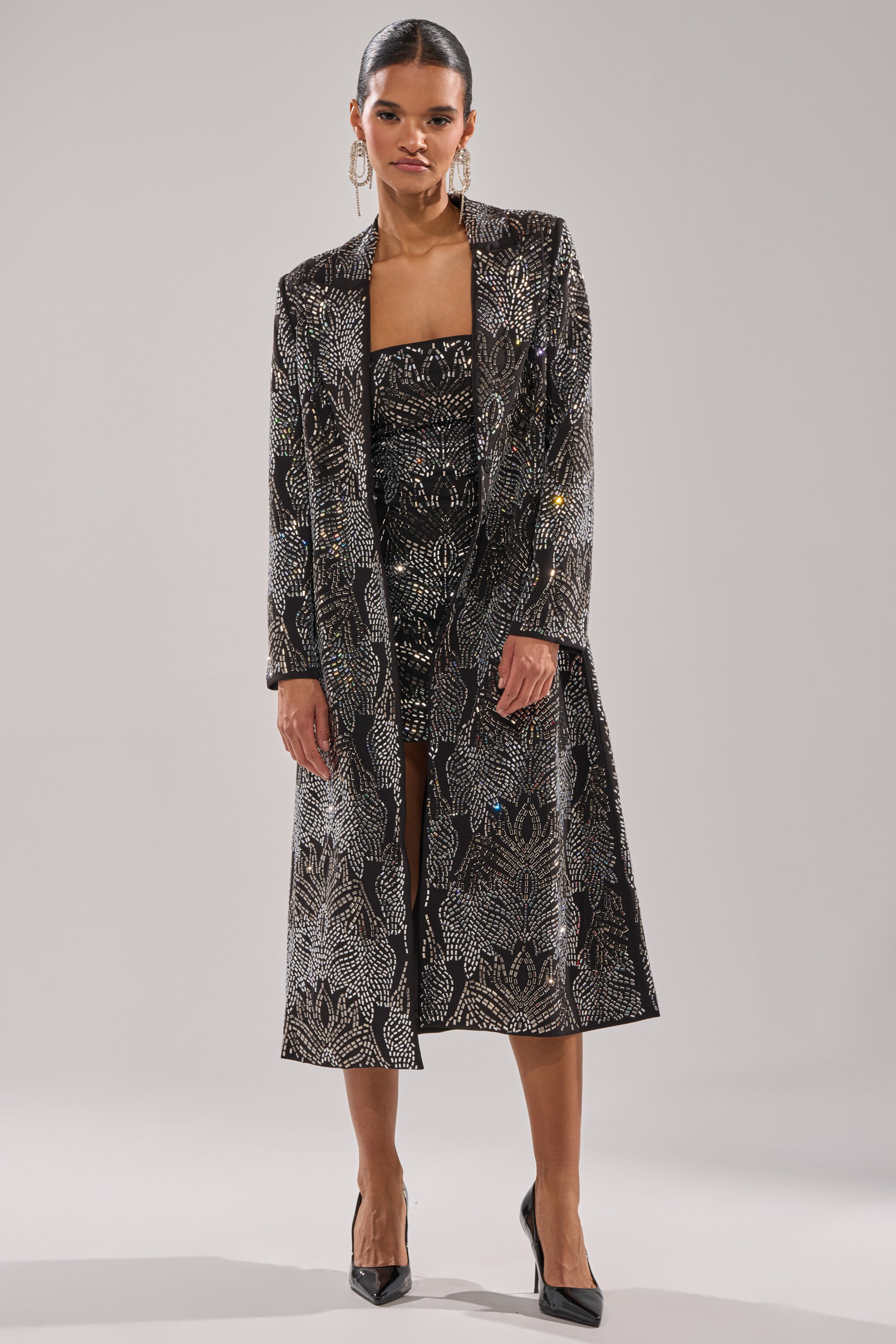 A woman stands facing forward against a plain background, wearing the SHE IS THE MOMENT RHINESTONE TRENCH over a matching fitted dress, paired with black pointed-toe heels and large hoop earrings. 5