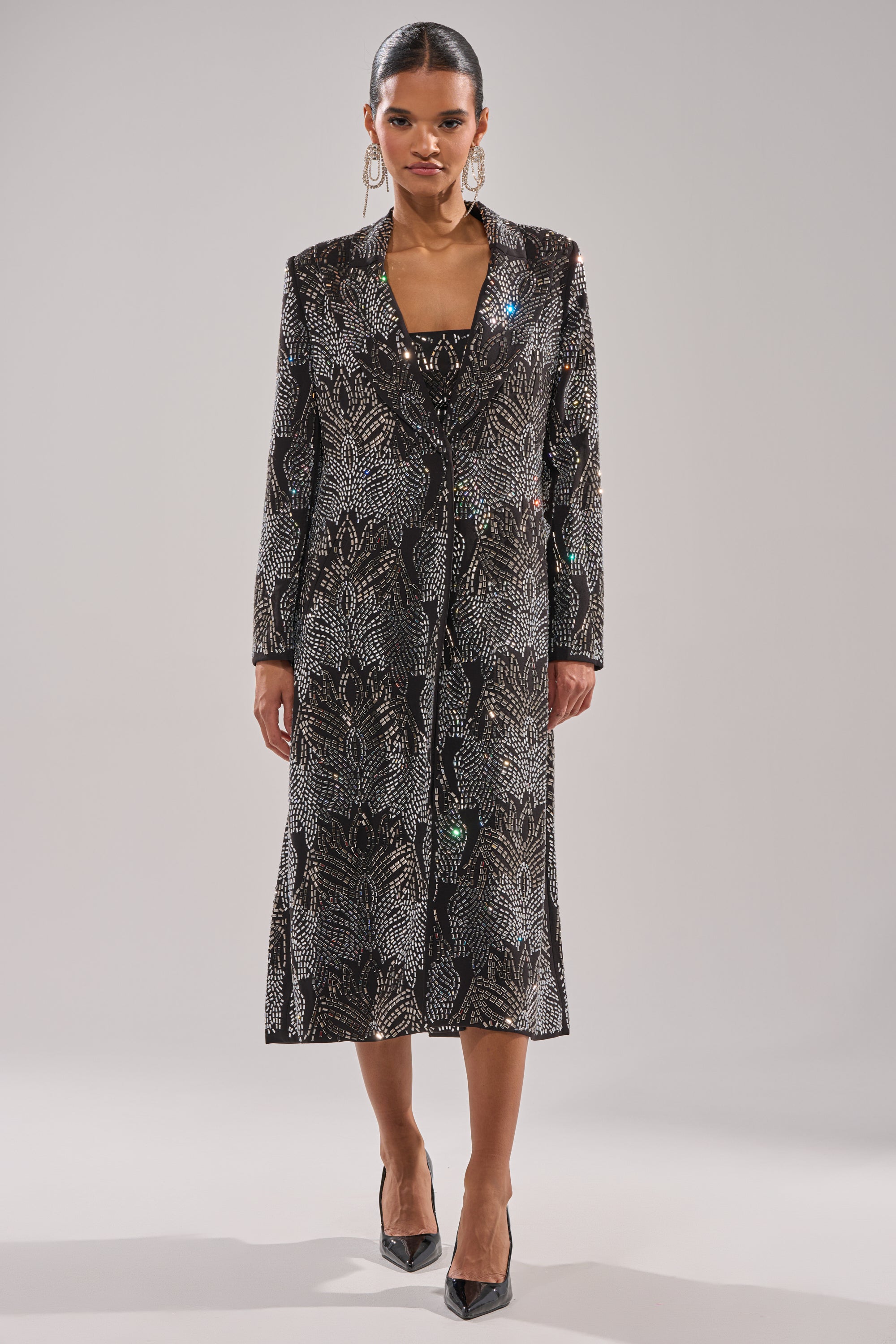 A woman stands facing forward in the SHE IS THE MOMENT RHINESTONE TRENCH, a longline coat dress with subtle sparkling details, paired with black pointed heels and large hoop earrings against a plain, light background. 7