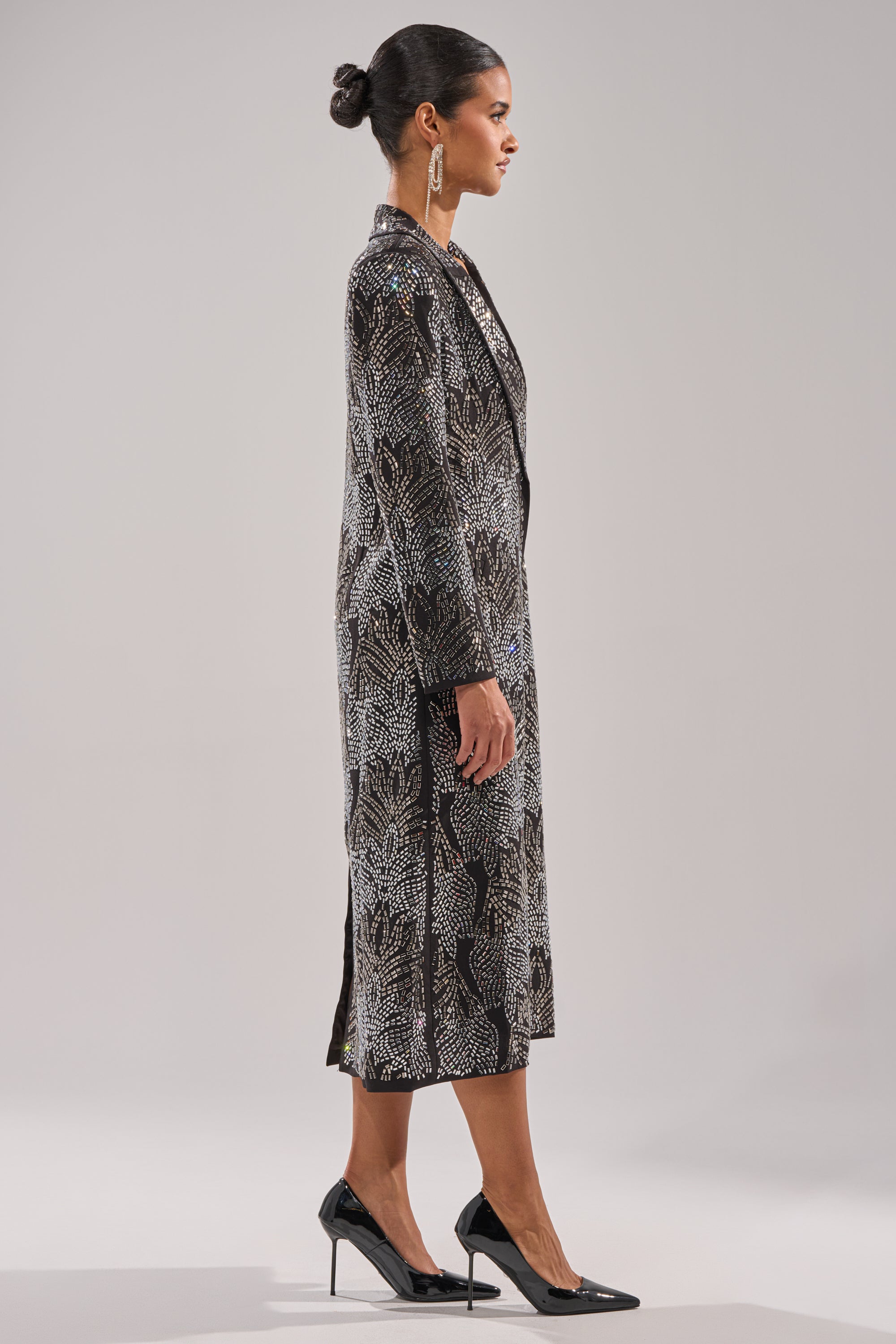 A woman in profile wears the SHE IS THE MOMENT RHINESTONE TRENCH, featuring a longline silhouette and black and white design, paired with black high heels and statement earrings. Her hair is in a neat bun against a light gray background. 8