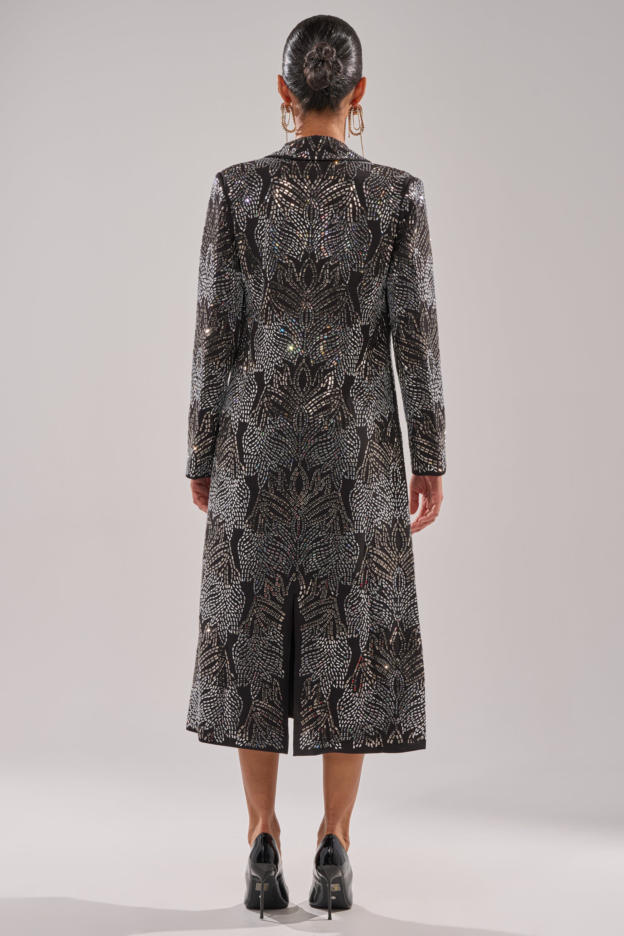 A person stands facing away in the SHE IS THE MOMENT RHINESTONE TRENCH, featuring a longline black silhouette with silver patterns. They wear black shoes, large hoop earrings, and have a sleek braided bun against a plain light gray backdrop. 9