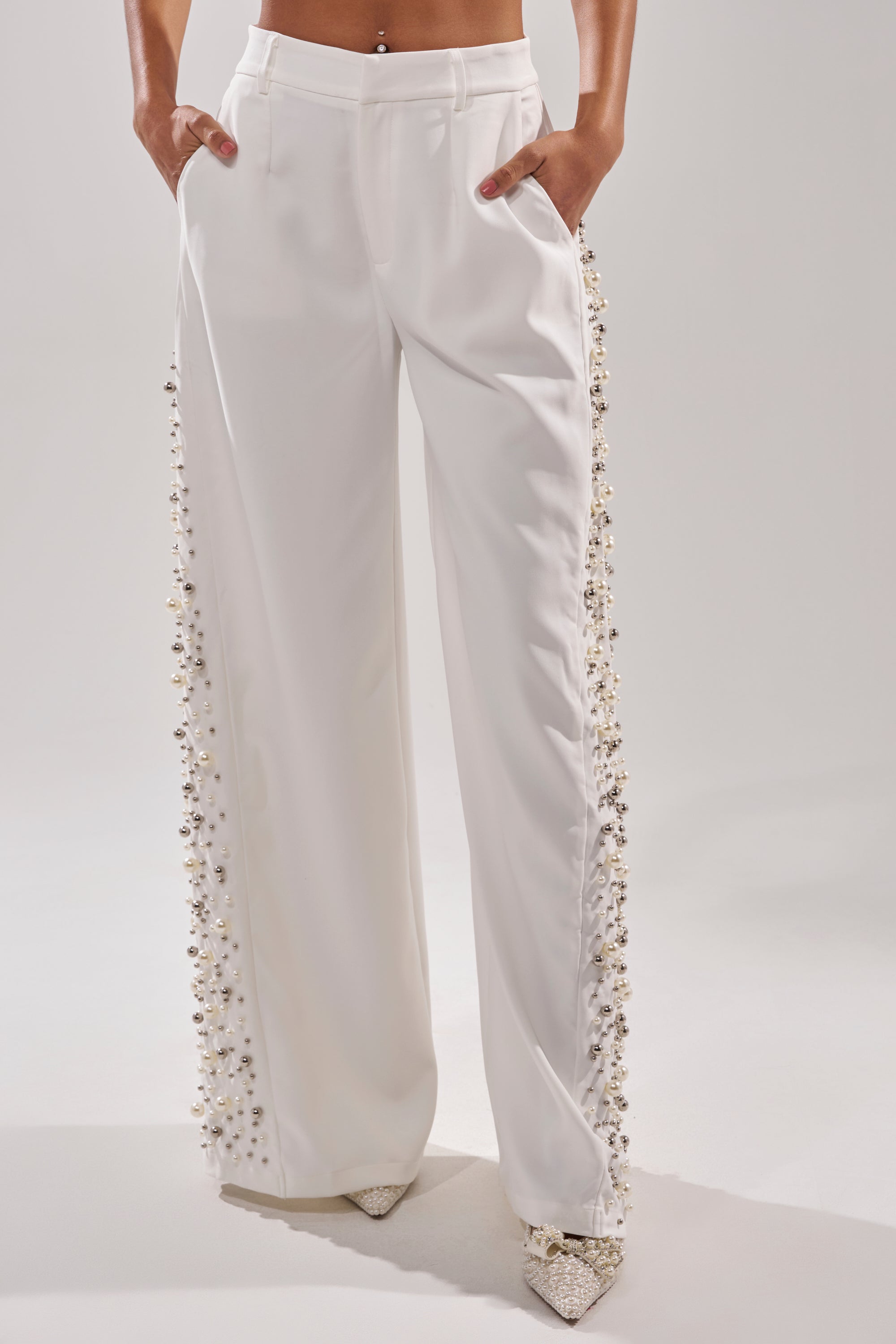 A person wears the KEEP IT GOING EMBELLISHED TROUSER—high-waisted, wide-leg pants with pearl and bead detailing along the side seams—hands in pockets and paired with pointed shoes.