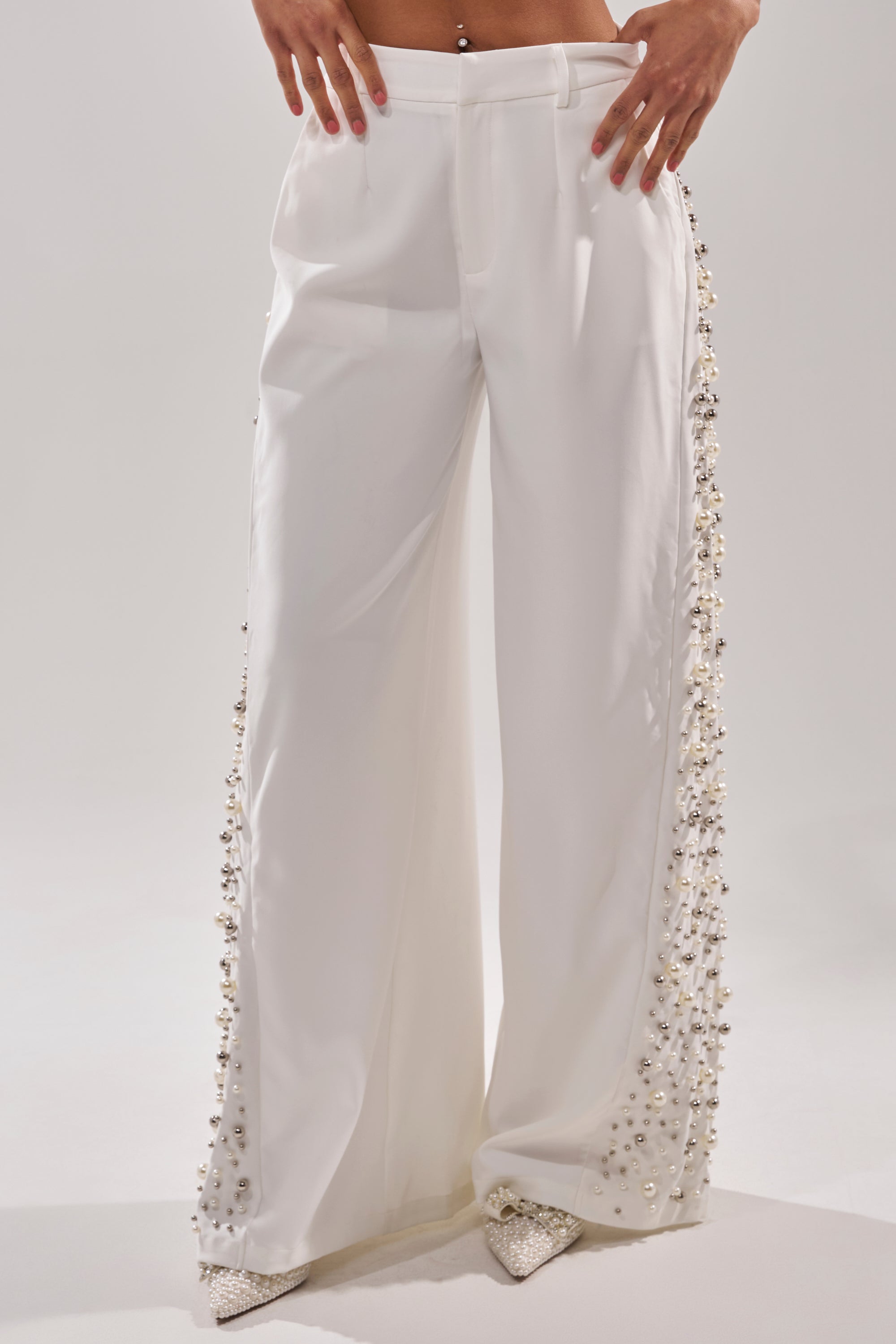 A person wearing the KEEP IT GOING EMBELLISHED TROUSER, featuring high-waisted, wide-leg pants with pearl embellishments along the side seams, paired with pointed-toe pearl shoes. Only their lower torso and legs are visible.