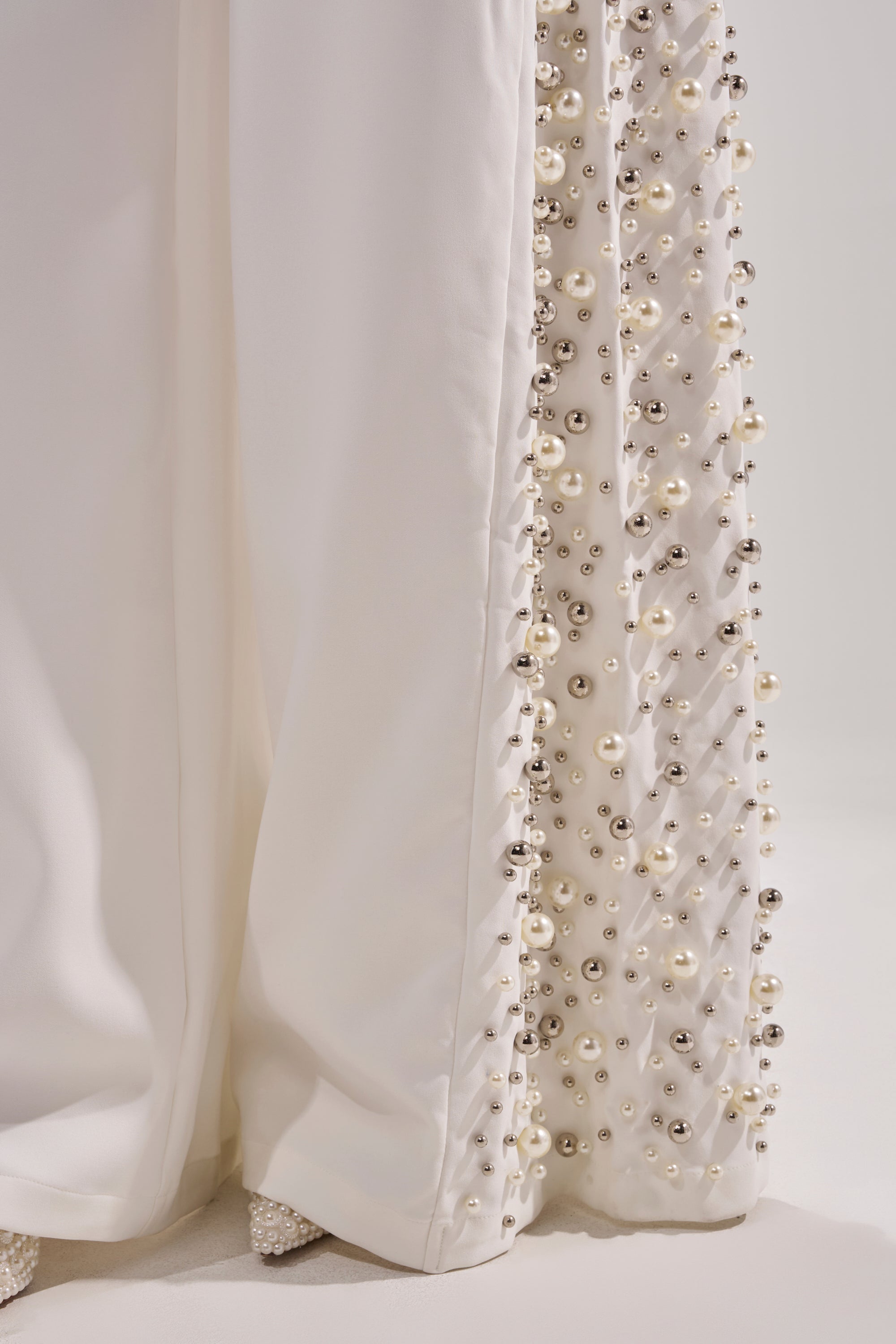 Close-up of the KEEP IT GOING EMBELLISHED TROUSER hem, showcasing rows of clustered pearls and silver studs densely decorating the edge, with fabric softly pooling on the floor.