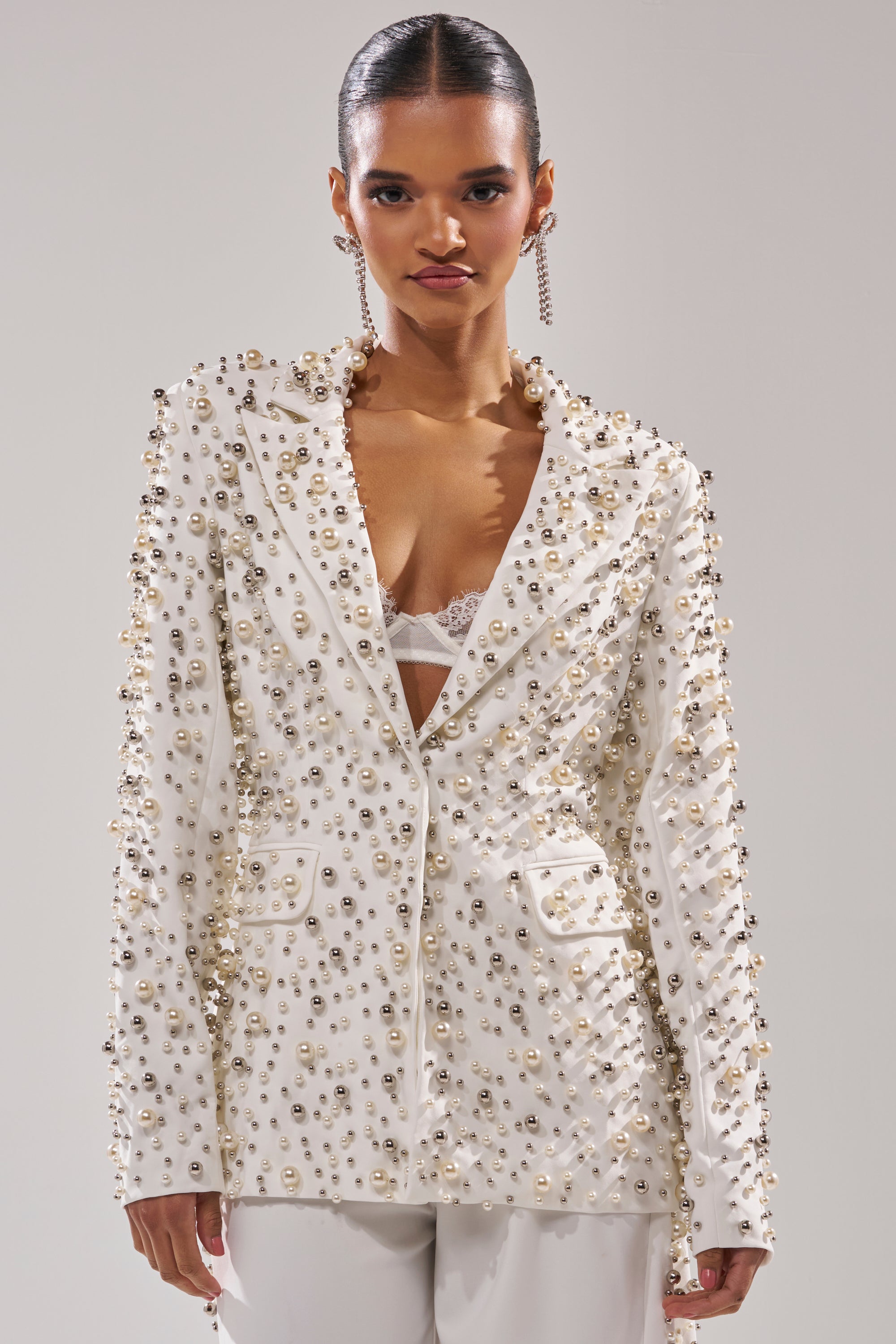 A woman with slicked-back hair models the KEEP IT GOING EMBELLISHED BLAZER with beaded pearls, paired with a white lace bra, white pants, and long earrings, set against a plain light background.