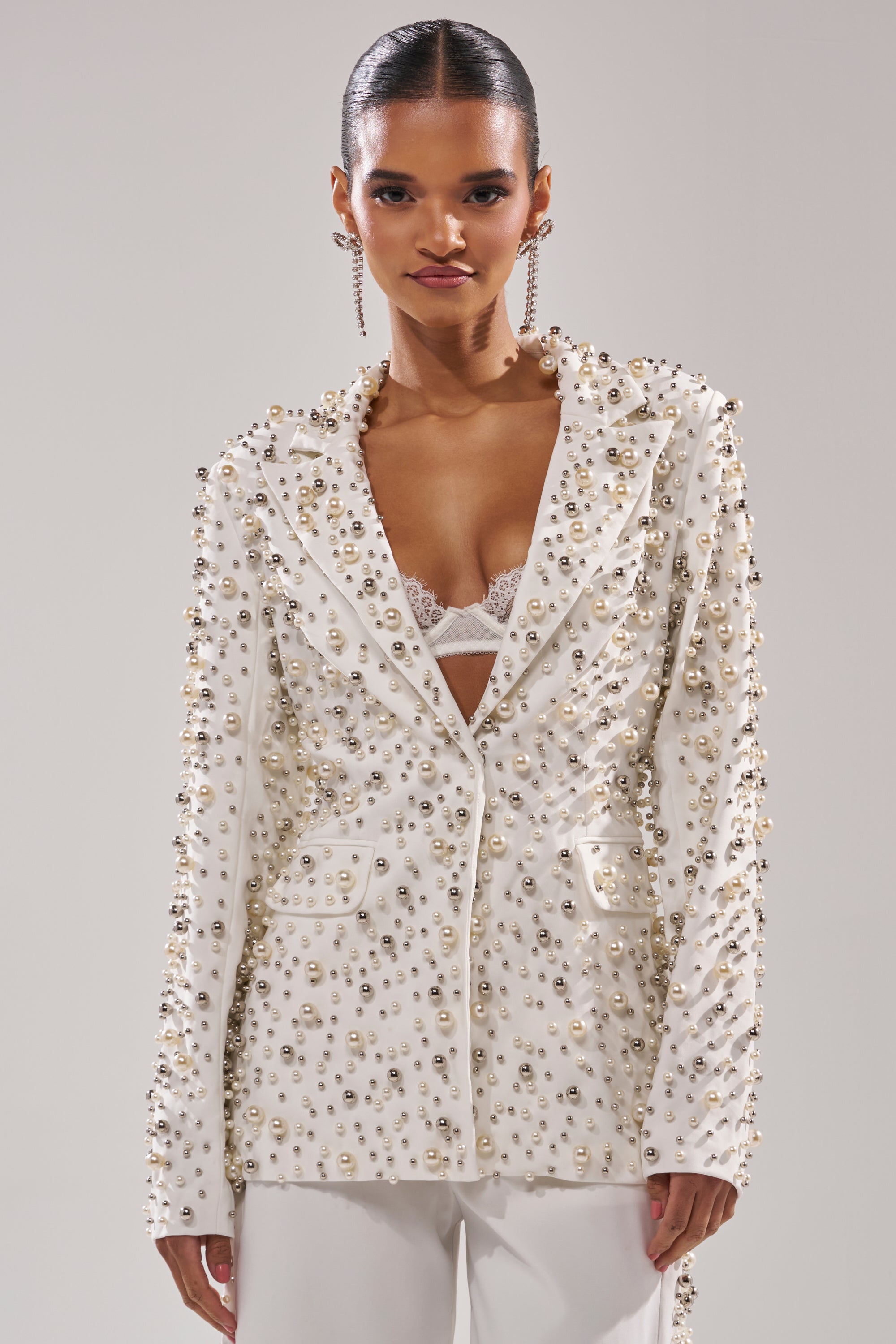 A woman styles the KEEP IT GOING EMBELLISHED BLAZER, adorned with metallic studs, over a white lace bra and pants. With slicked-back hair and long dangling earrings, she creates a chic, edgy look.
