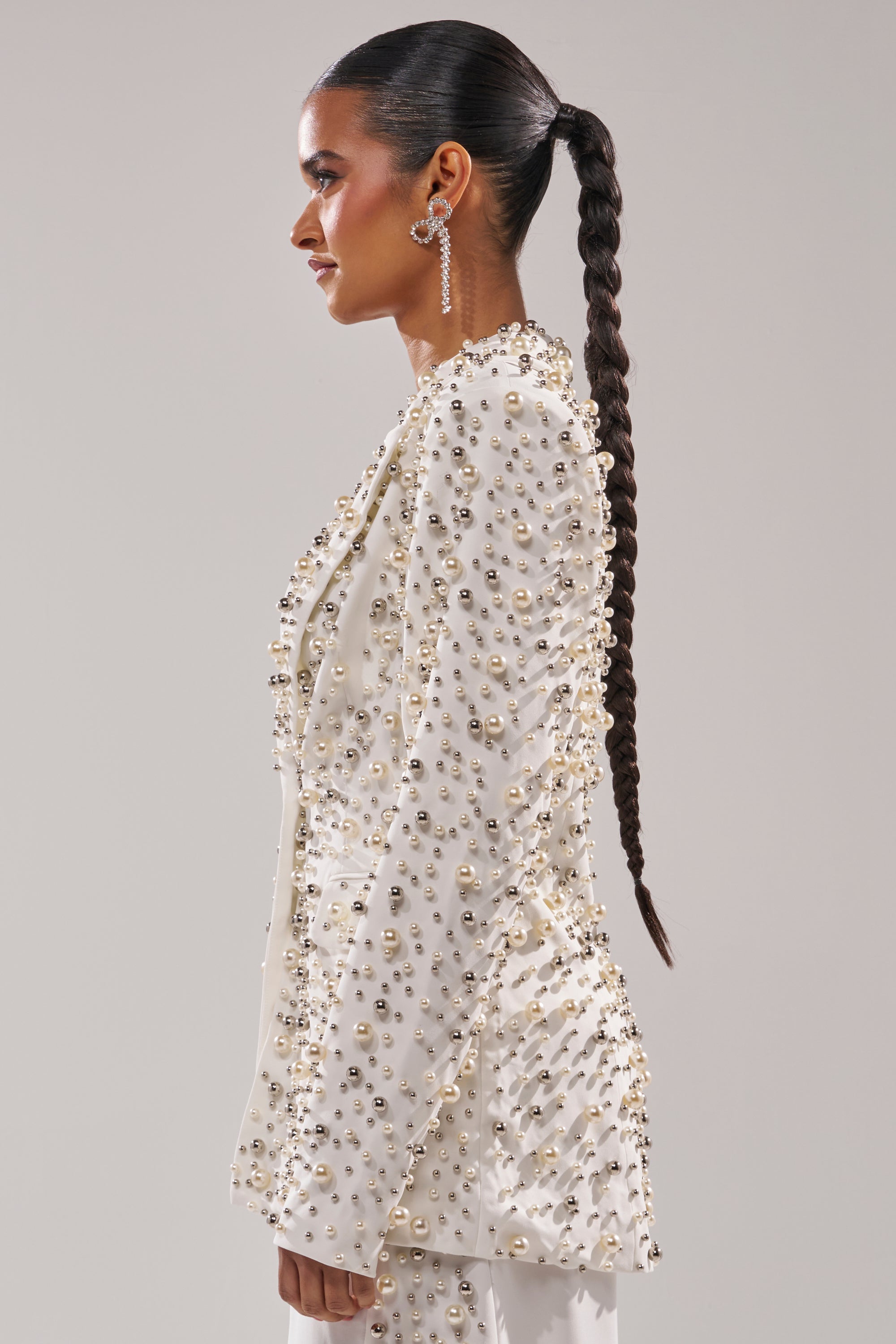 A woman with a long braided ponytail is seen in profile wearing the KEEP IT GOING EMBELLISHED BLAZER, featuring pearl beading and a tailored silhouette. Statement earrings and metallic details complete her polished, elegant look.