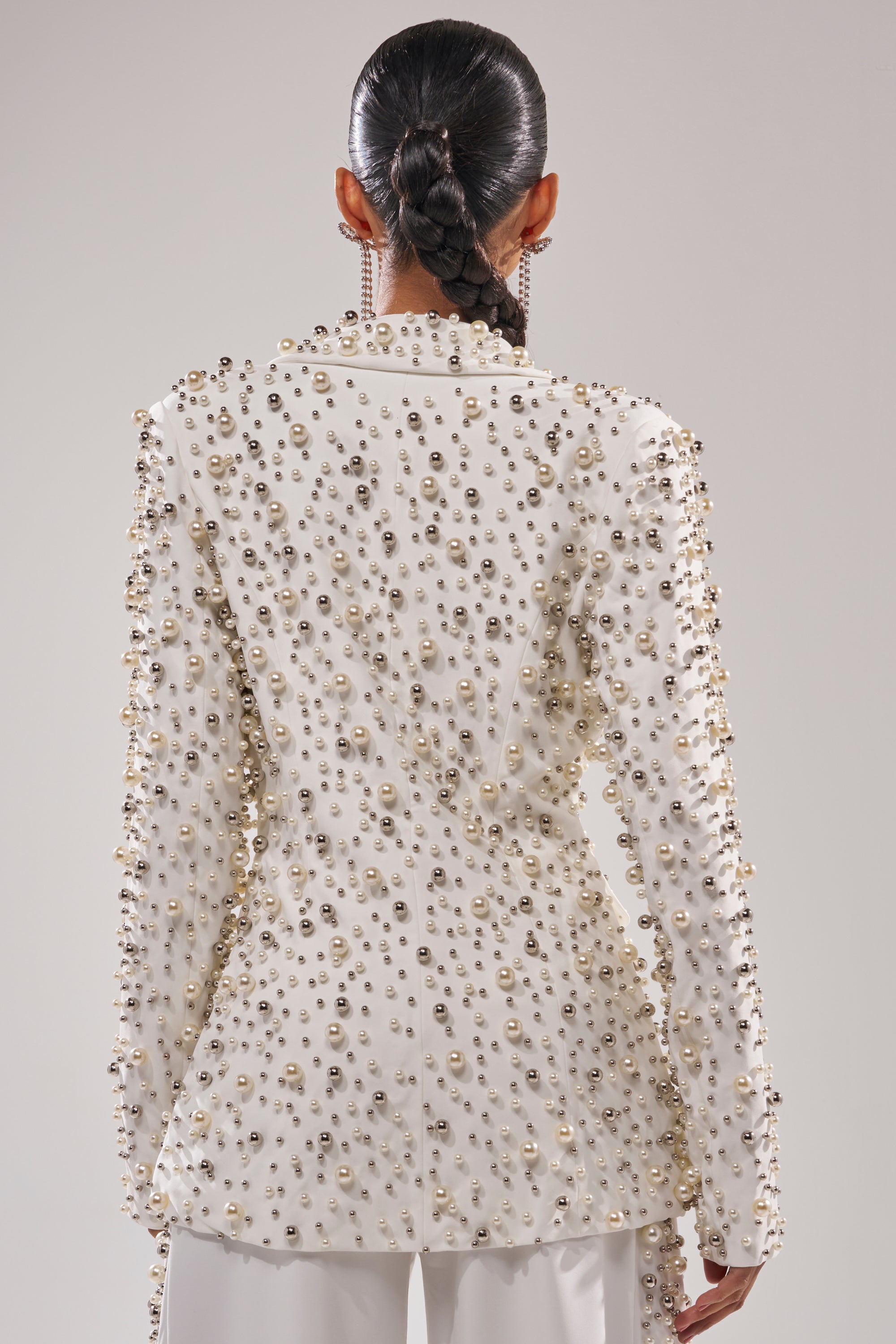 A person with dark, slicked-back braided hair is seen from behind in the KEEP IT GOING EMBELLISHED BLAZER, adorned with pearls and metallic studs, paired with matching white pants for a bold tailored look against a plain background.